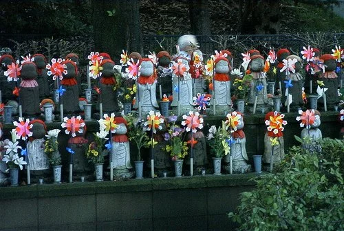 A collection of small garden decorations featuring Jizo figures wearing hats and scarves, each adorned with colorful pinwheels, placed among potted plants and flowers on a raised stone platform outdoors.