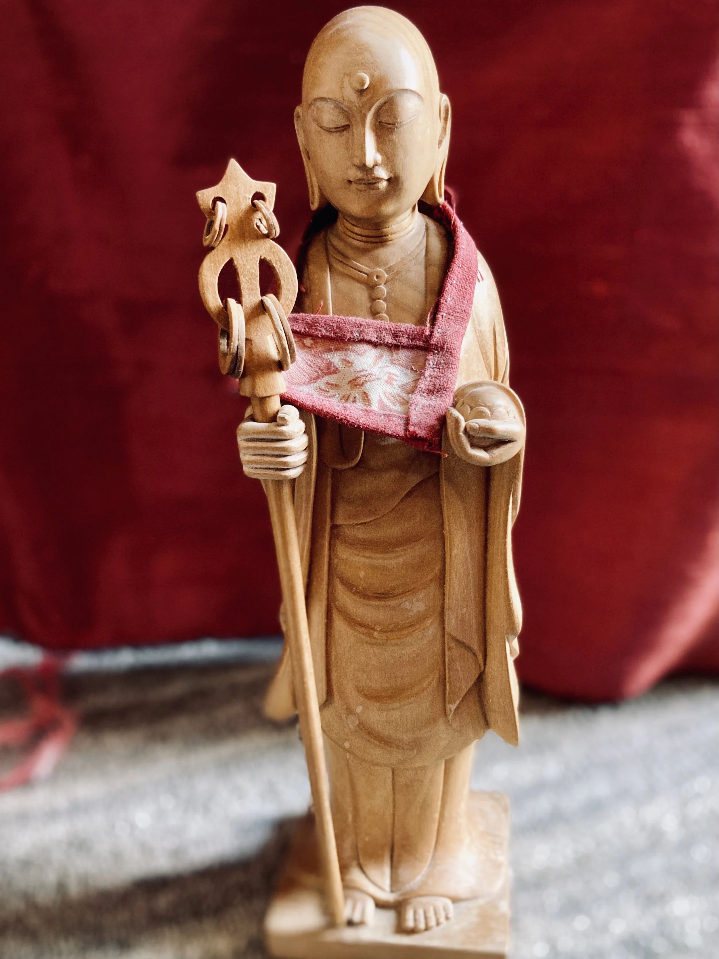 Wooden figurine of a serene, meditative person with closed eyes, holding a staff in the left hand and a bowl in the right, dressed in traditional attire with jewelry, standing against a red background.