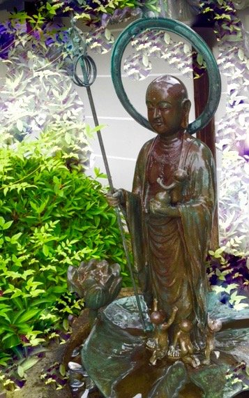 Bronze statue of a child monk holding a staff with a circular ring, standing on a base surrounded by green and purple plants.