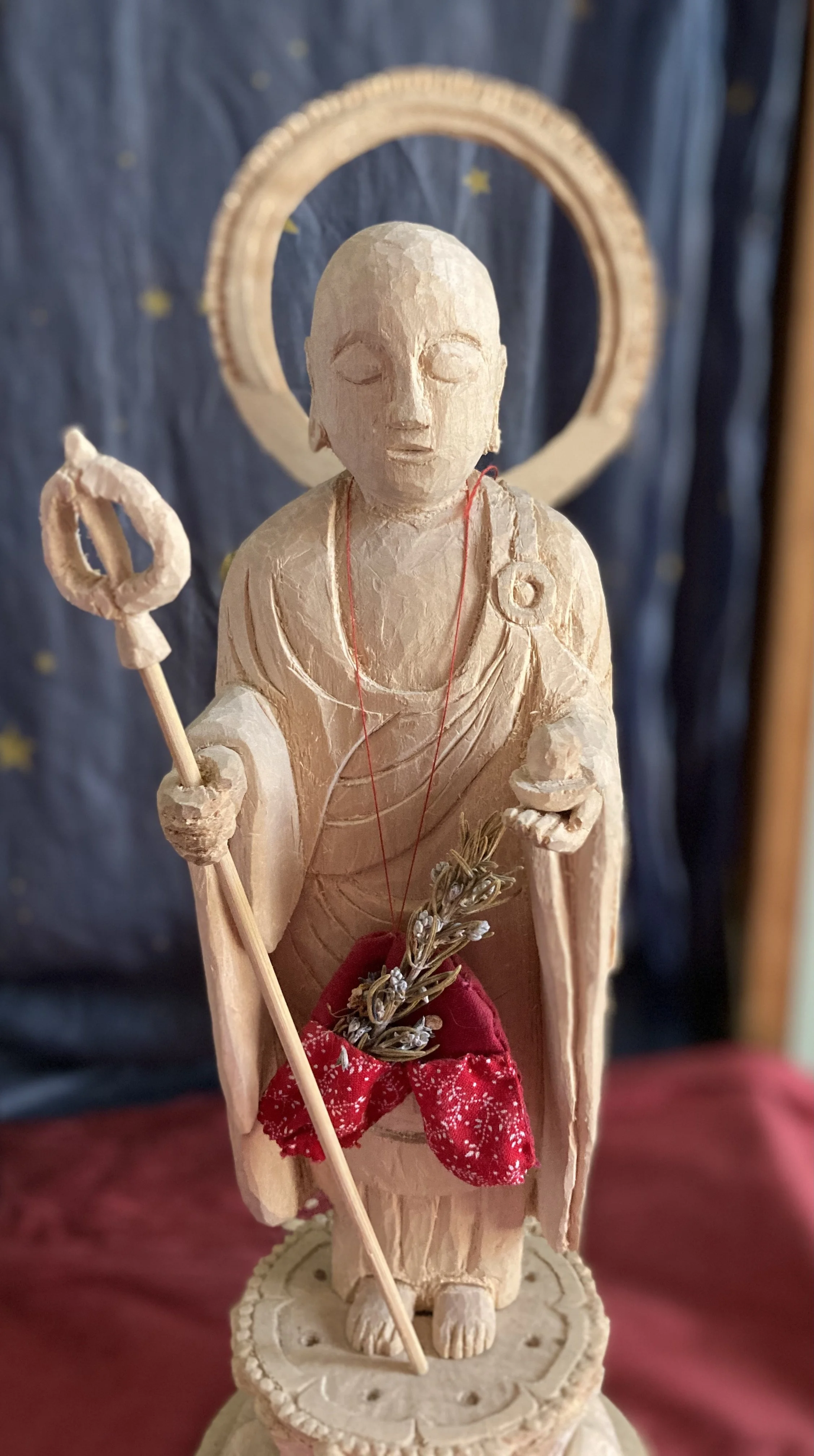 Wooden statue of a person with closed eyes, holding a staff in the right hand and a small branch in the left, dressed with a red cloth, standing on a decorative base with a halo behind head, against a backdrop of dark blue fabric with yellow stars.