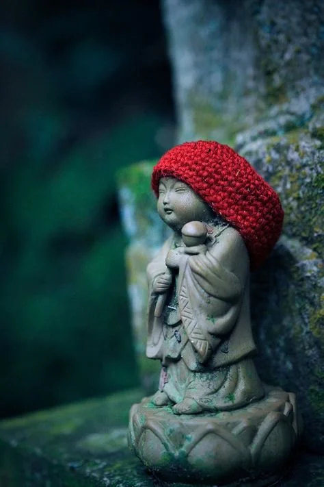 Small stone statue of a Buddhist monk with a red knit hat, holding a staff, standing against a mossy rock wall.