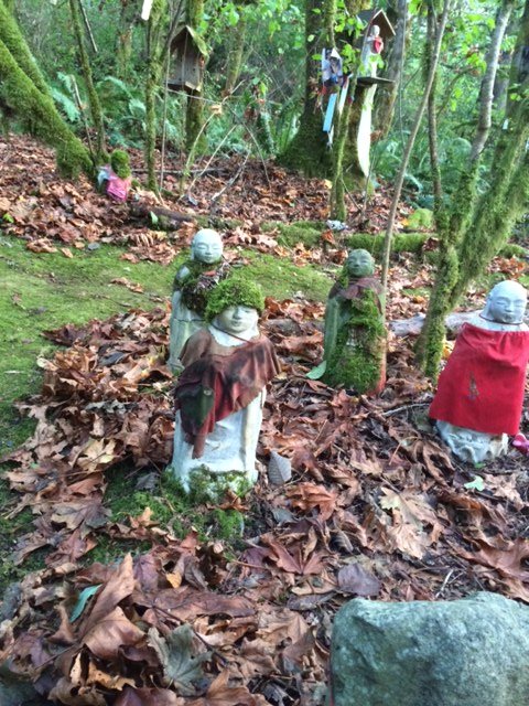 Multiple small Jizo stone statues in a forest, some covered with moss and cloth, amidst fallen leaves and trees.