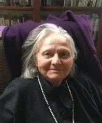 An elderly woman with gray hair, wearing a black top and a pearl necklace, sitting in a chair in a room with bookshelves in the background.