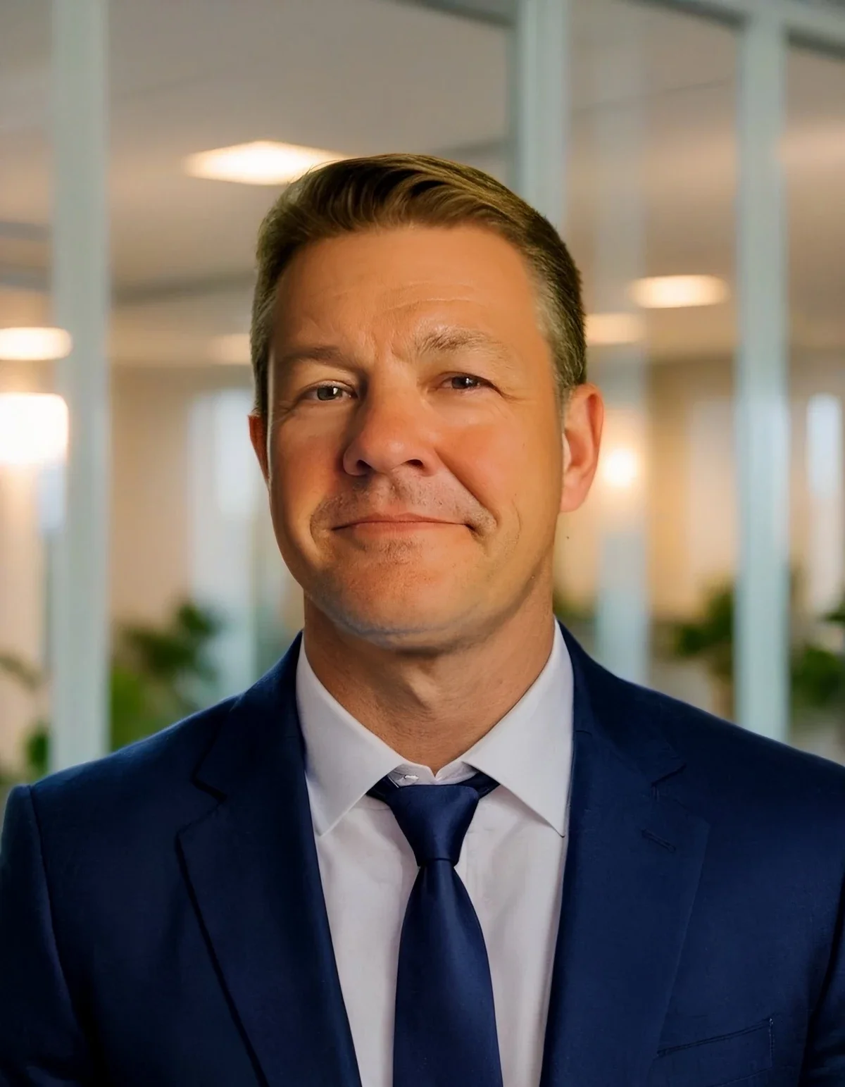 Professional man wearing a dark blue suit, white shirt, and navy tie standing in a modern office with glass walls and blurred background lights.