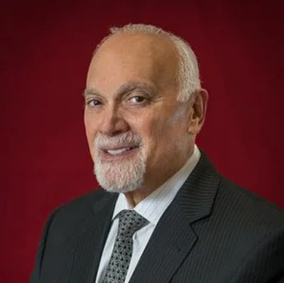 Portrait of an older man with a bald head, gray beard, wearing a dark suit, white shirt, and patterned tie, against a red background.
