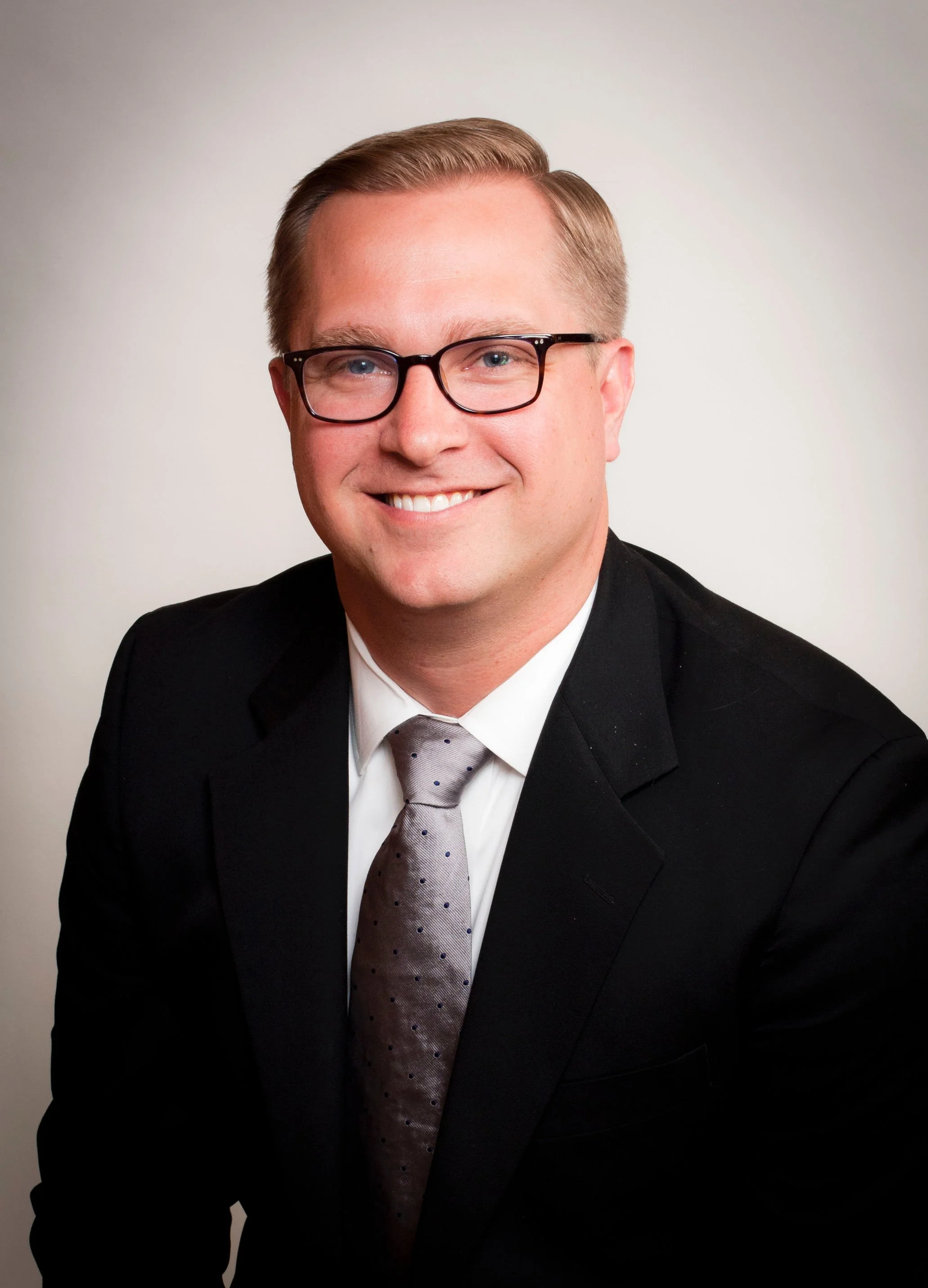 Professional portrait of a man wearing glasses, a dark suit, white shirt, and a patterned gray tie, smiling against a plain, light background. Scott Baltsen COO