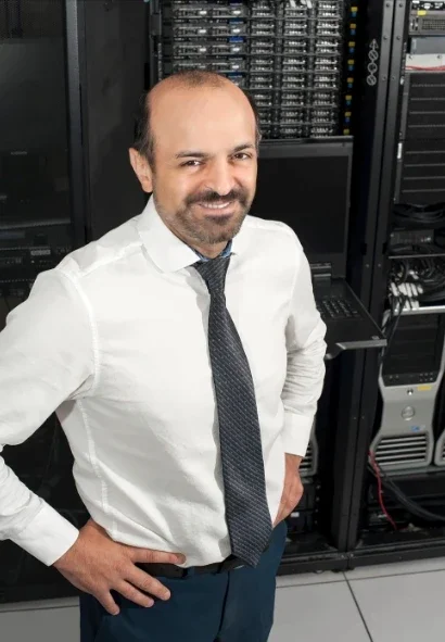 A man in a white dress shirt and tie standing in front of server racks in a data center or server room.