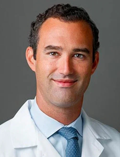 A male doctor with dark hair, wearing a white lab coat and a light blue tie, smiling against a gray background.