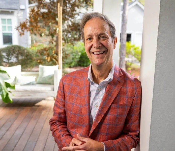 A smiling man in a red plaid blazer standing on a porch with greenery in the background. Dr. Jim Robinson Founder and President