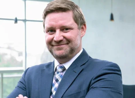 A man in a suit smiling with arms crossed in a modern office setting.