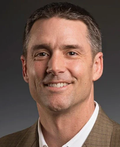 Professional headshot of a man with short brown hair and a light beard, wearing a suit jacket and white shirt, smiling against a dark background.