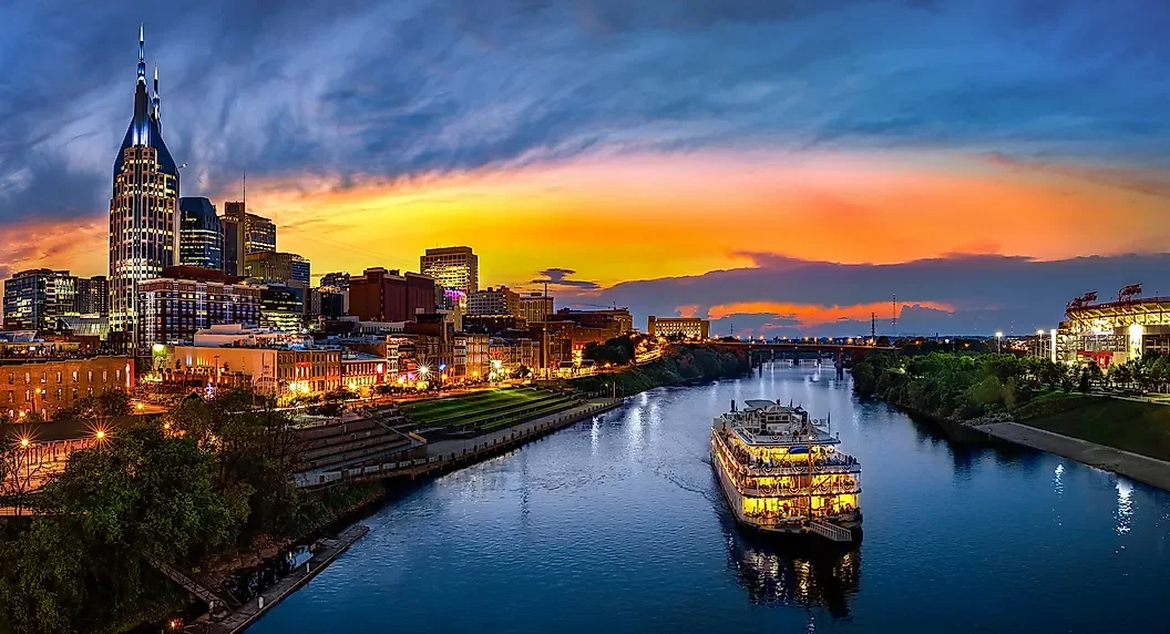 A city skyline at sunset with a river in the foreground, a cruise boat on the river, and tall skyscrapers including the AT&T building in Nashville, Tennessee.