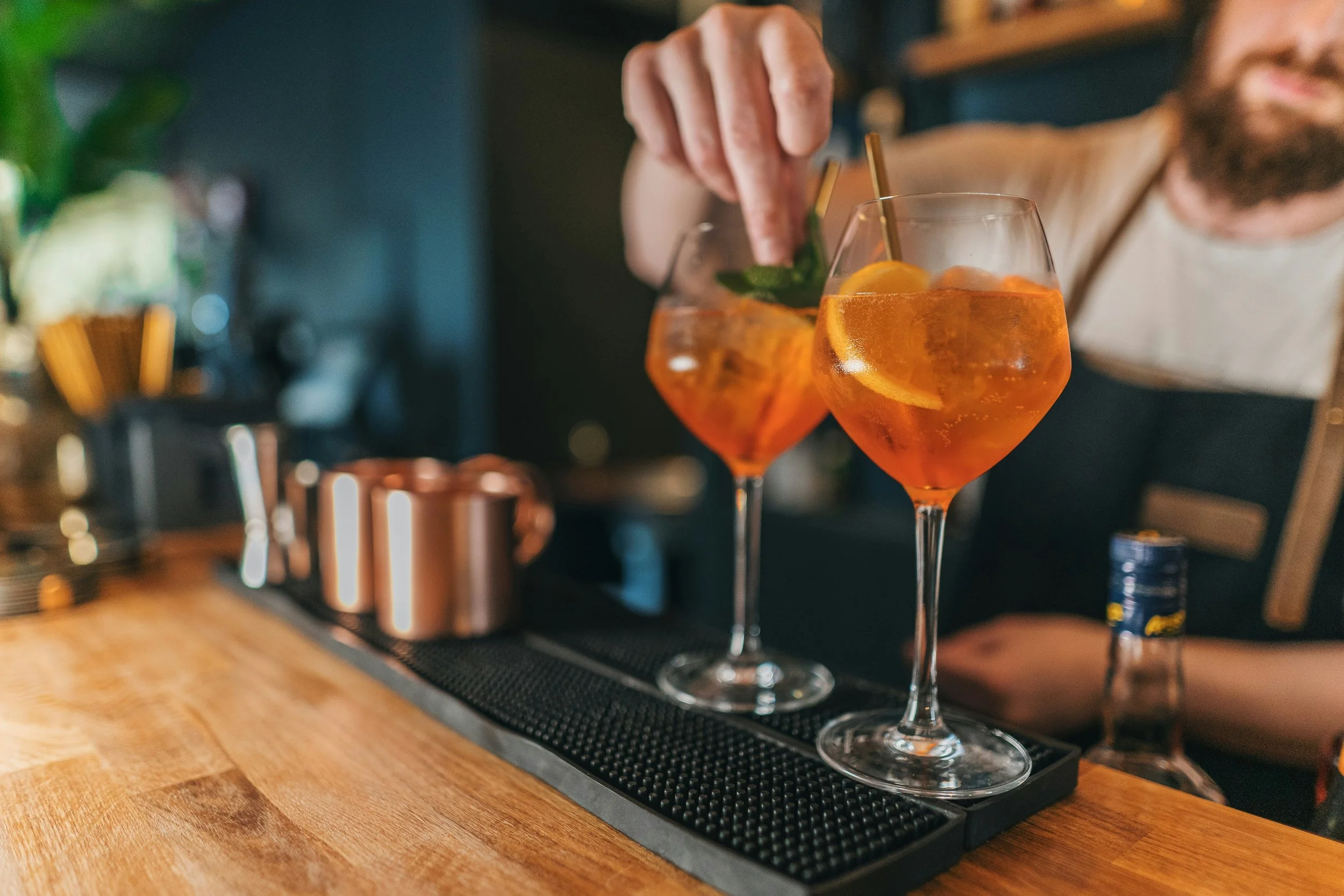 Bartender preparing two Aperol spritz cocktails with orange slices and mint leaves on a bar counter.