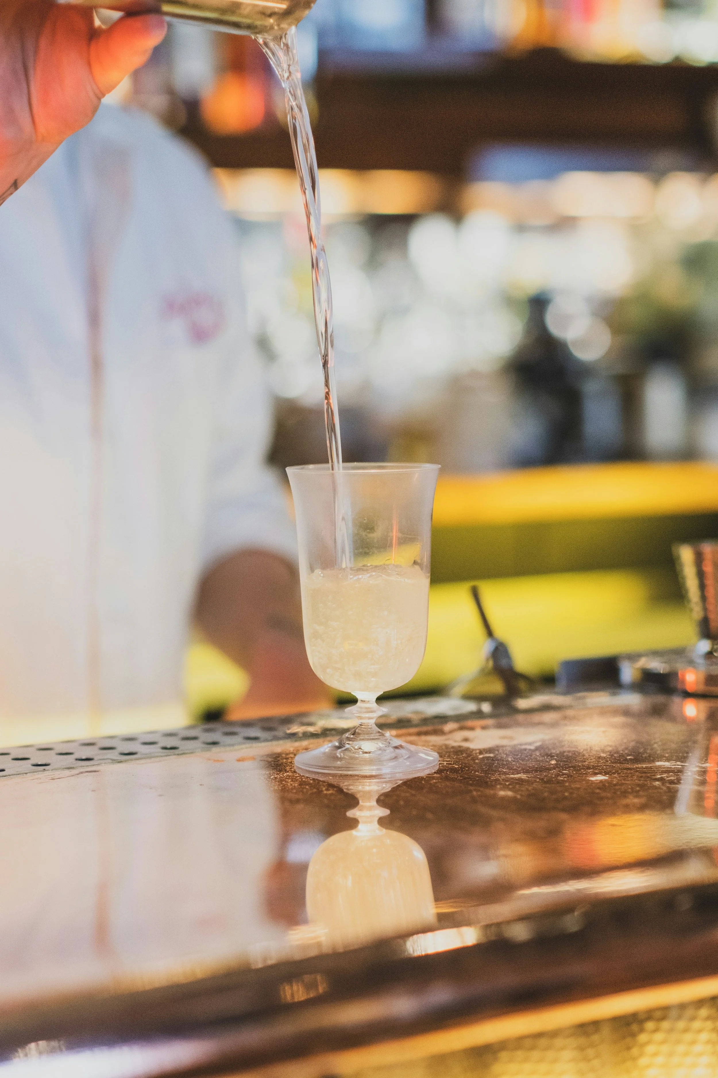 A person pouring a clear carbonated beverage from a glass into a cocktail glass on a bar counter.