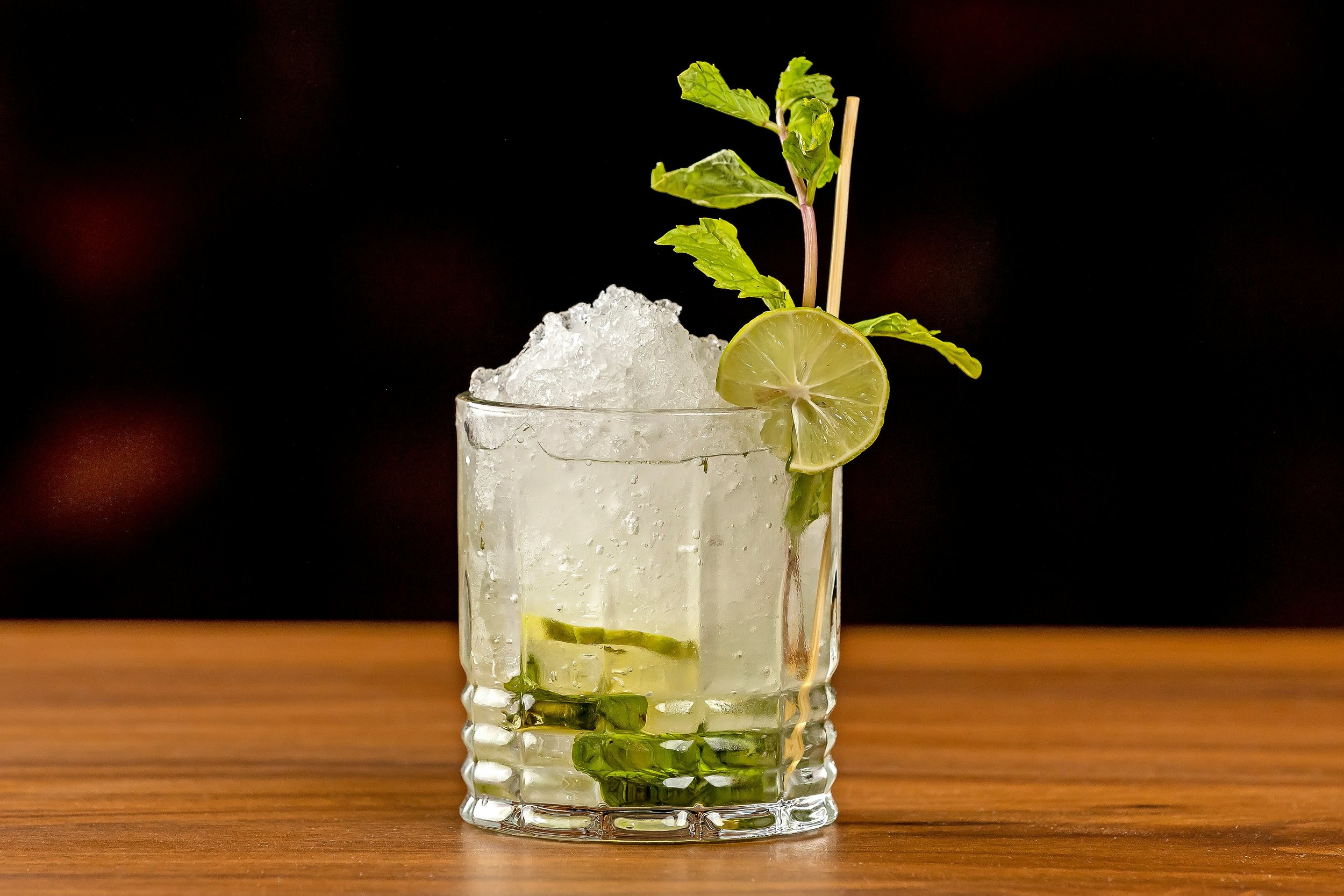 A glass of mojito with crushed ice, lime slices, mint leaves, and a straw on a wooden surface with a dark background.