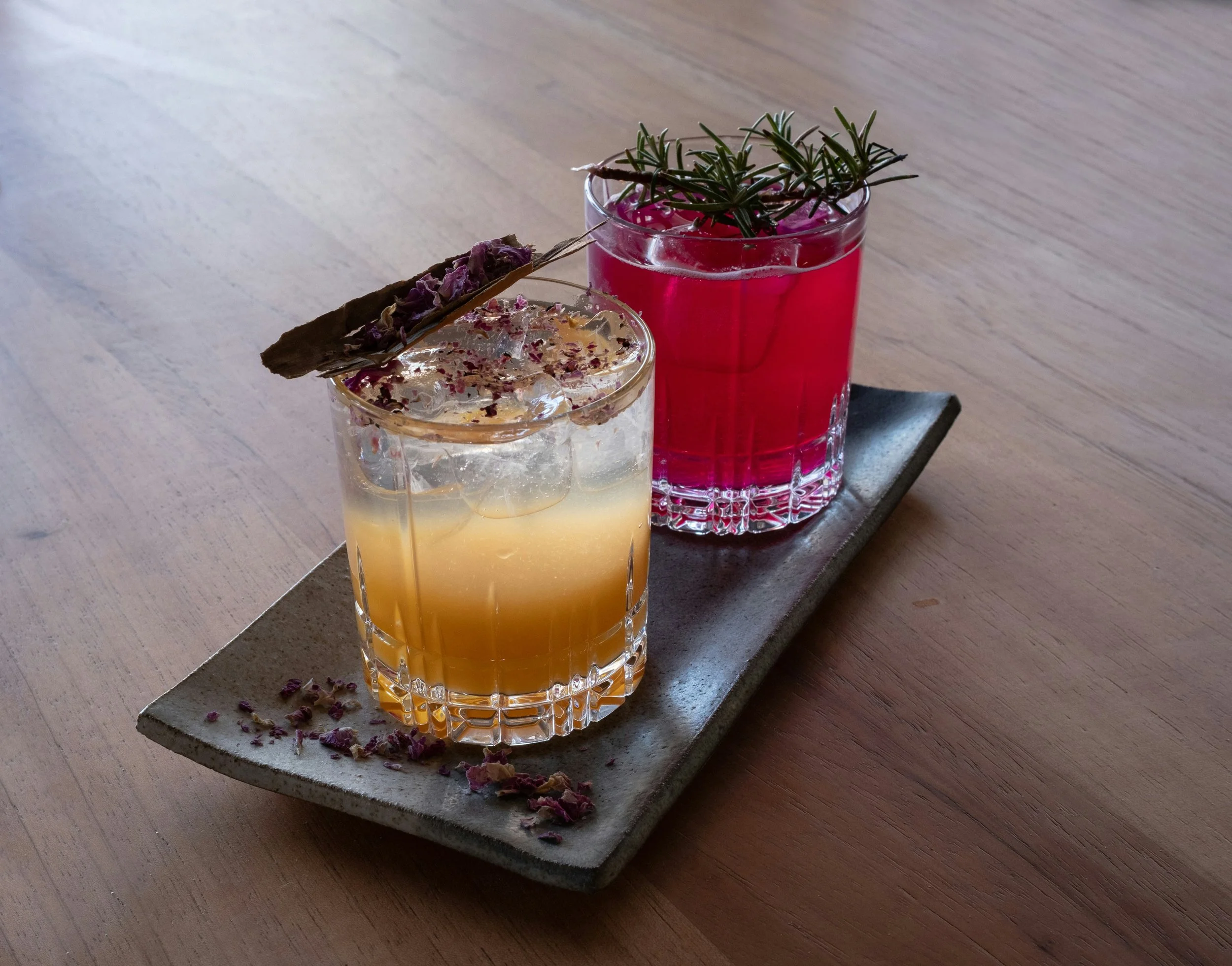 Two colorful cocktails on a rectangular ceramic plate with flowers for garnish, placed on a wooden table.