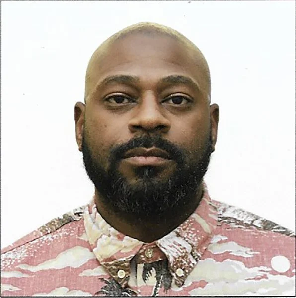 A close-up portrait of a man with a beard and short, light-colored hair. He is wearing a patterned, button-up shirt and has a neutral expression against a plain background.