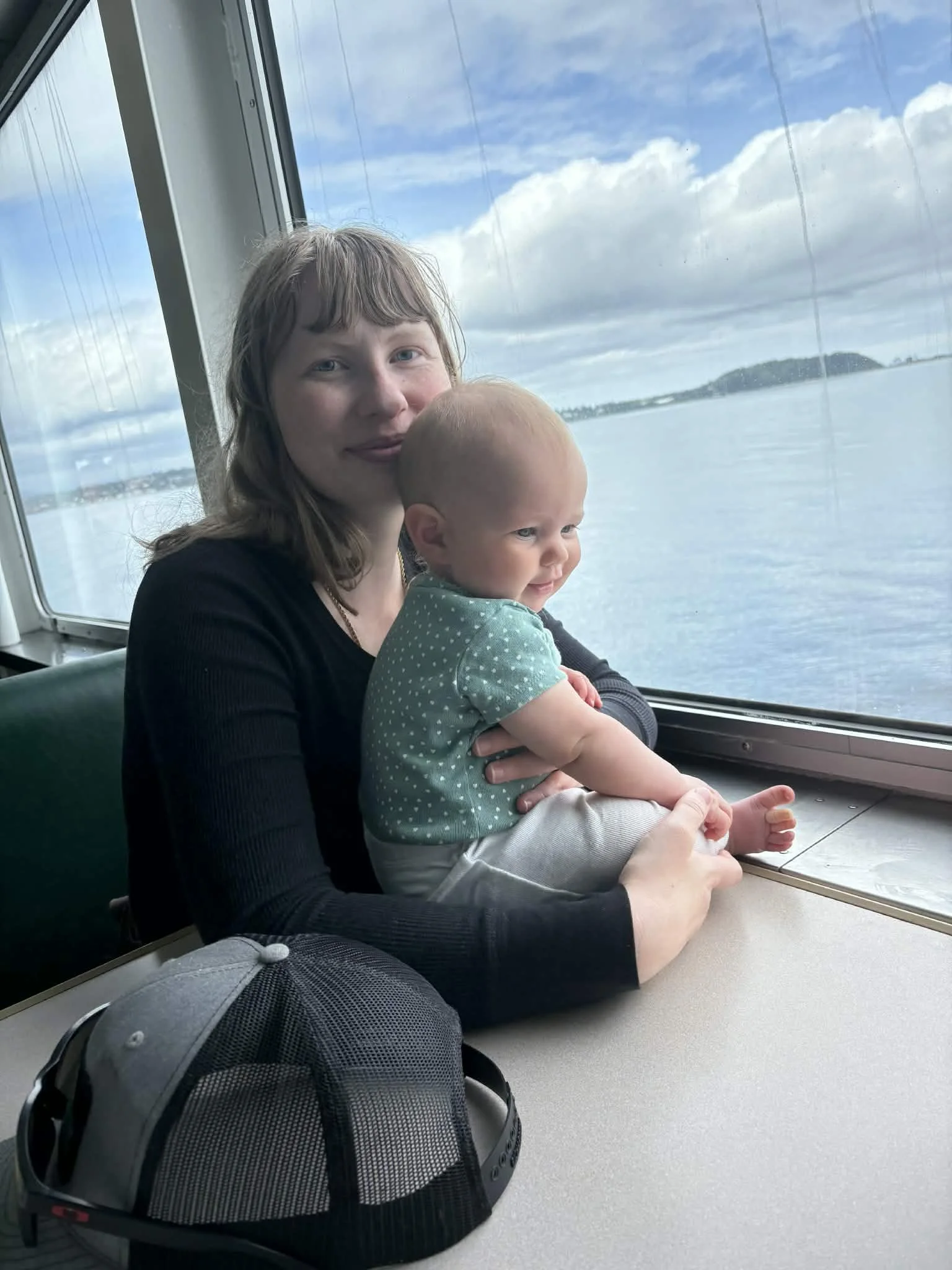 mother and baby sitting in a booth on a ferry. clouds, ocean, sky, and islands in the distance.
