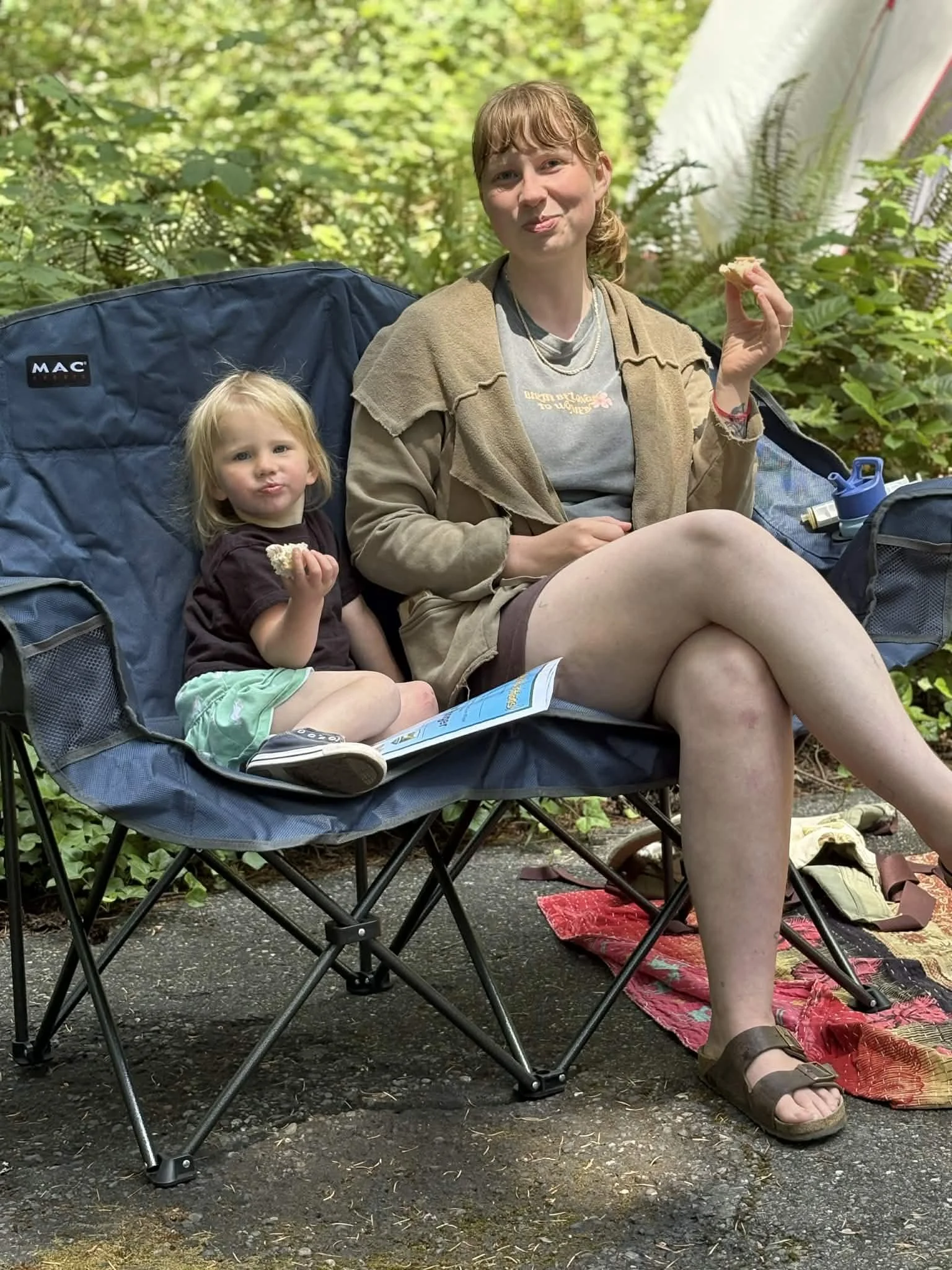 mother and toddler sitting in a camping chair with food in hand. they are in a green forest
