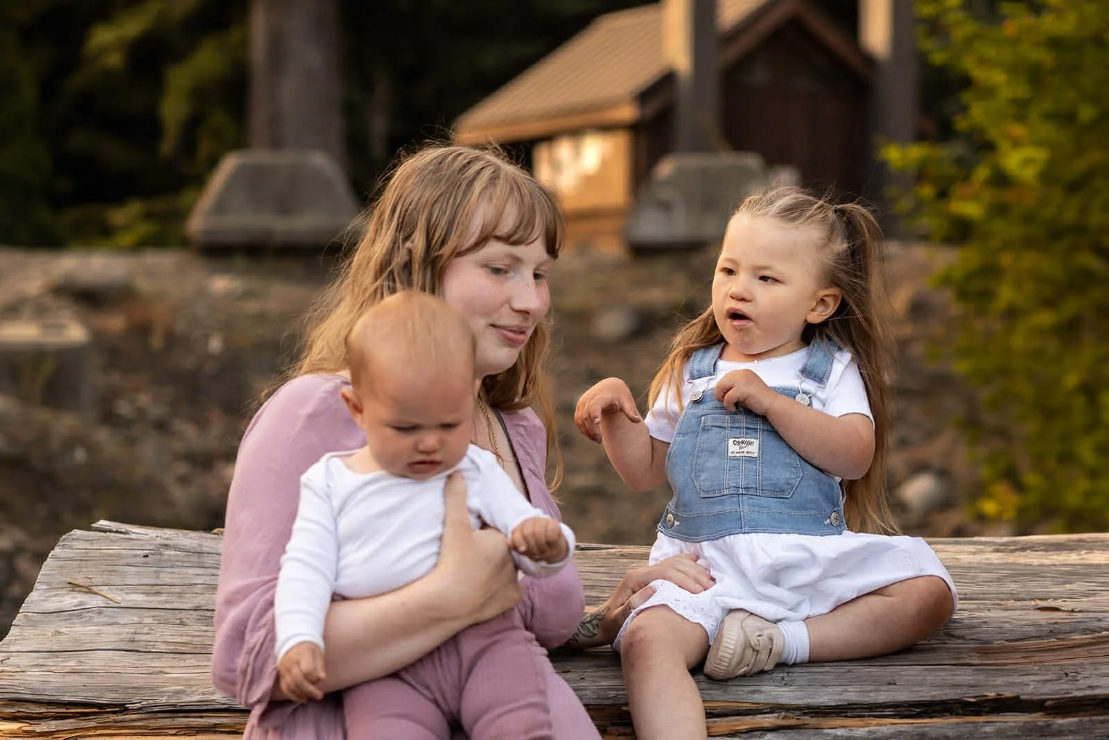 mother and two daughters, one sitting on a large fallen tree. at a lake with a building in the background.