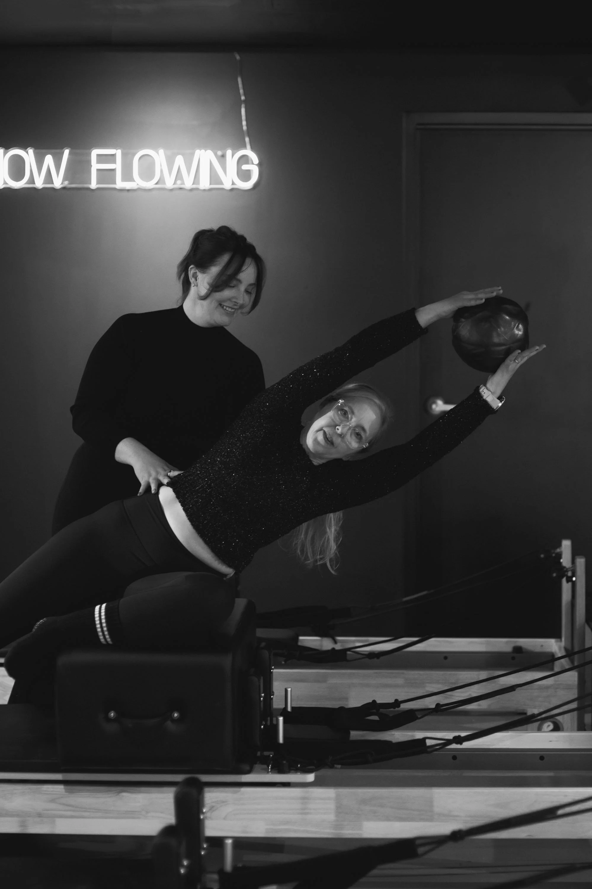 Two women in a fitness studio demonstrating or practicing Pilates or a similar workout; one woman is lying on a reformer machine holding a ball overhead, while the other supports her; neon sign reads 'NOW FLOWING' in the background.