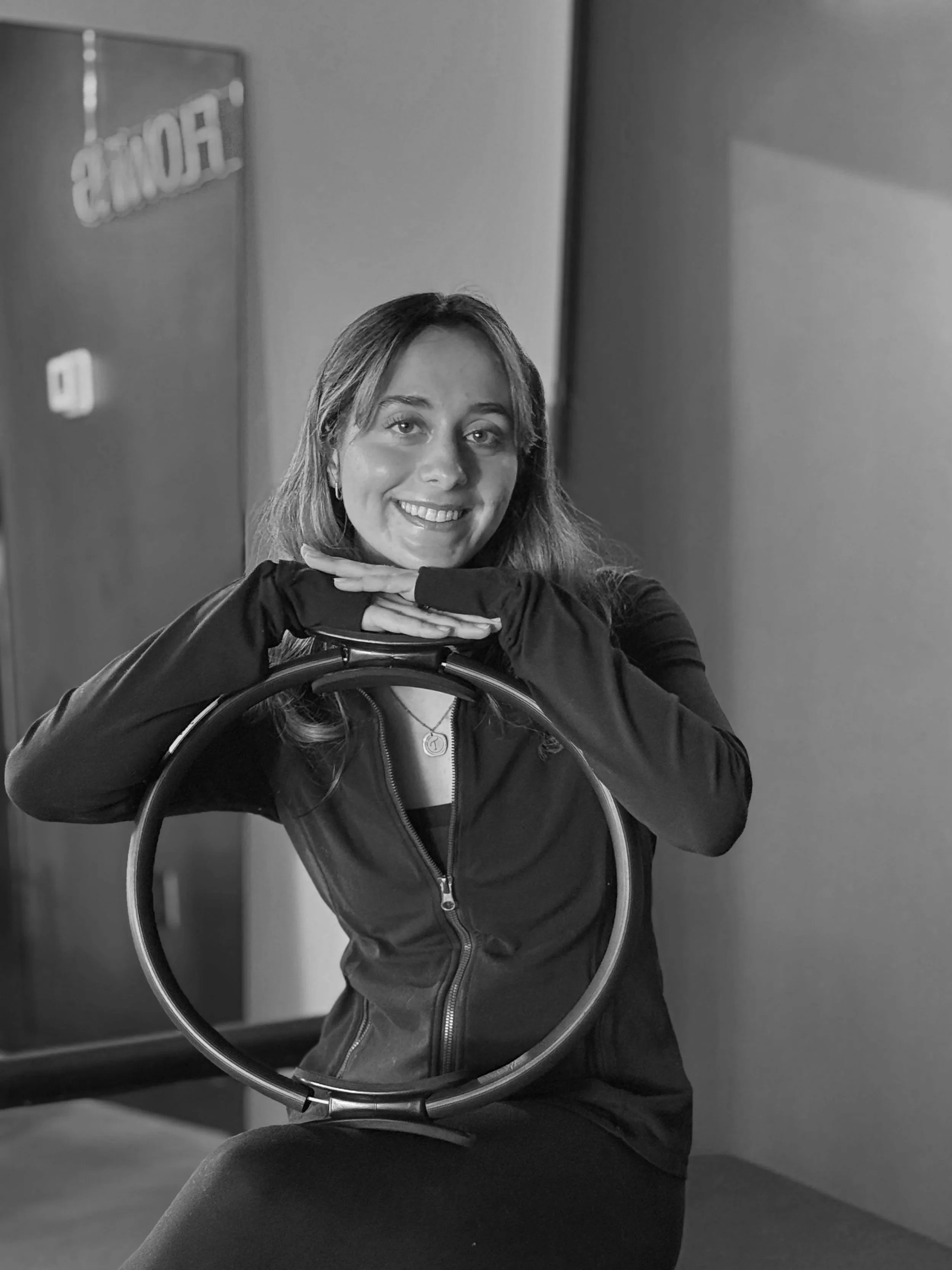A woman with short hair, wearing a black long-sleeve top, sitting on a Pilates reformer machine in a fitness studio with neon signs reading 'NOW FLOW' and 'NOW FLOW' in the background.