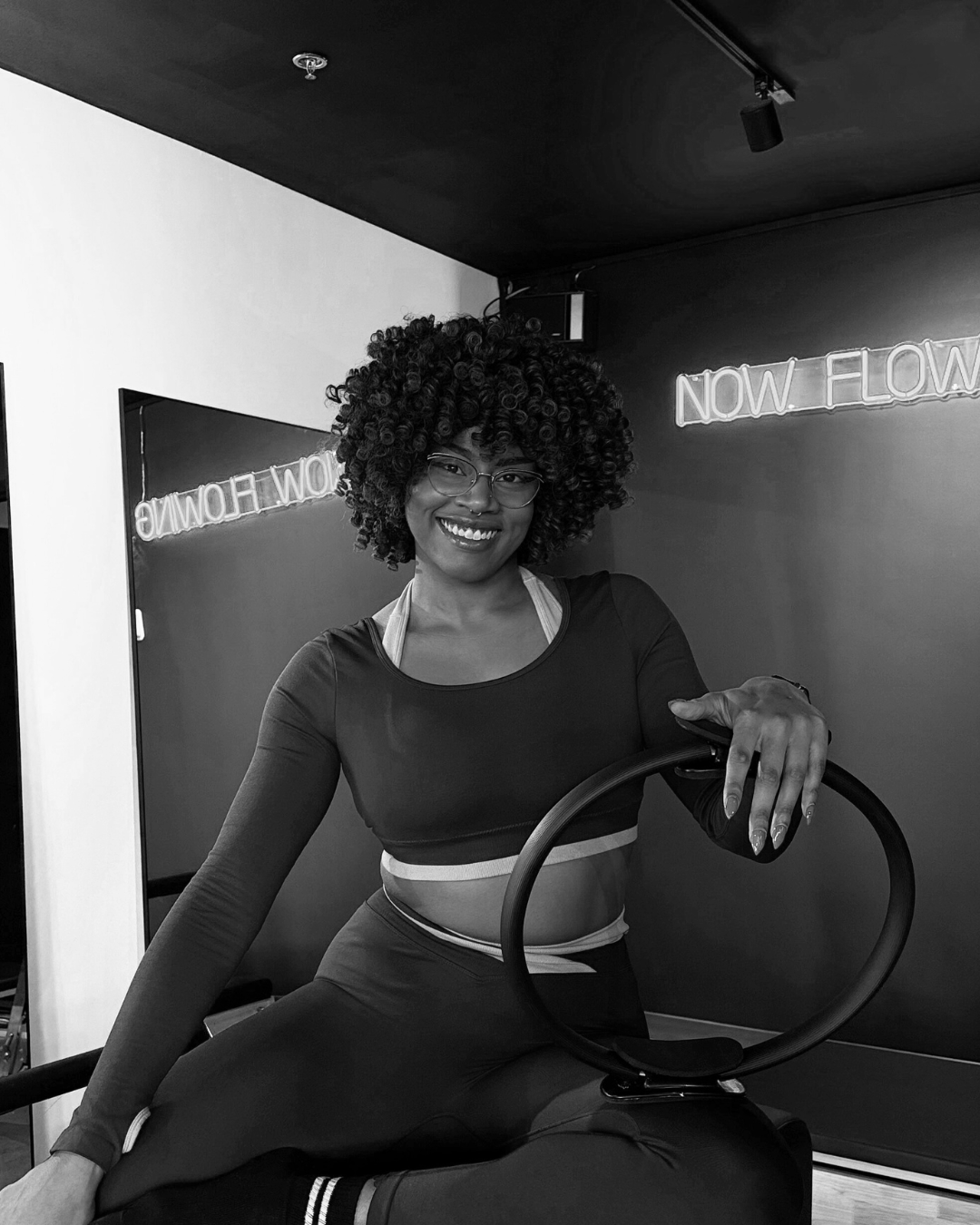 A woman with curly hair and glasses smiling while sitting on a fitness equipment, in a gym with neon signs in the background.