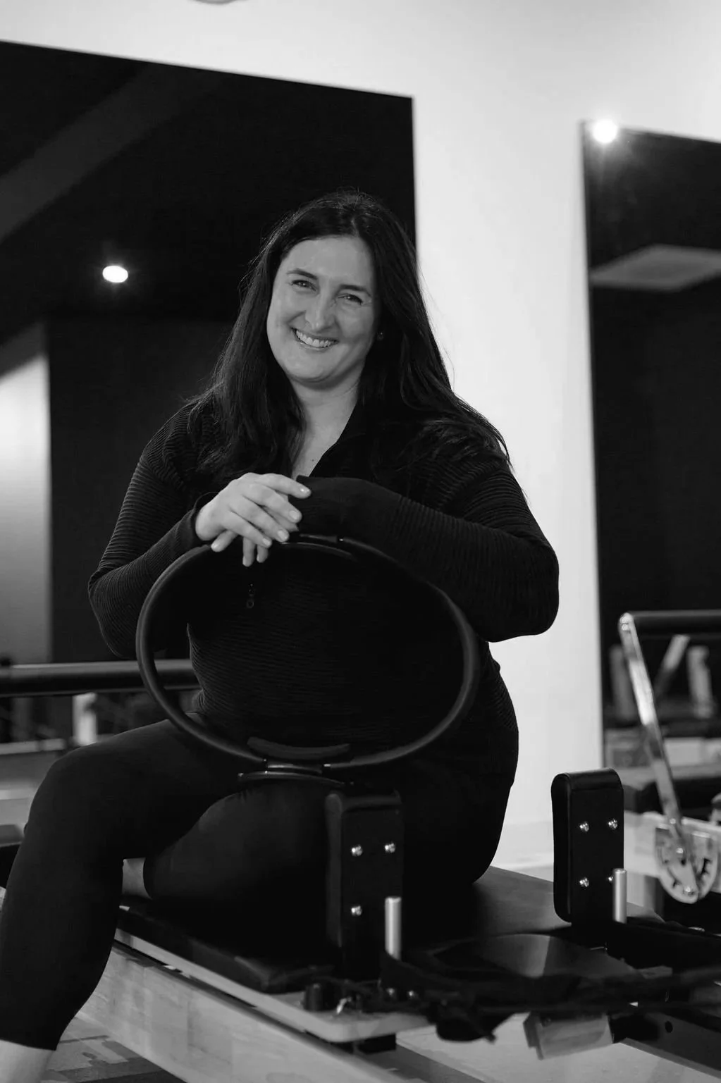 A woman with long dark hair smiling, sitting on an exercise Pilates reformer machine in a fitness studio, in black and white photo.