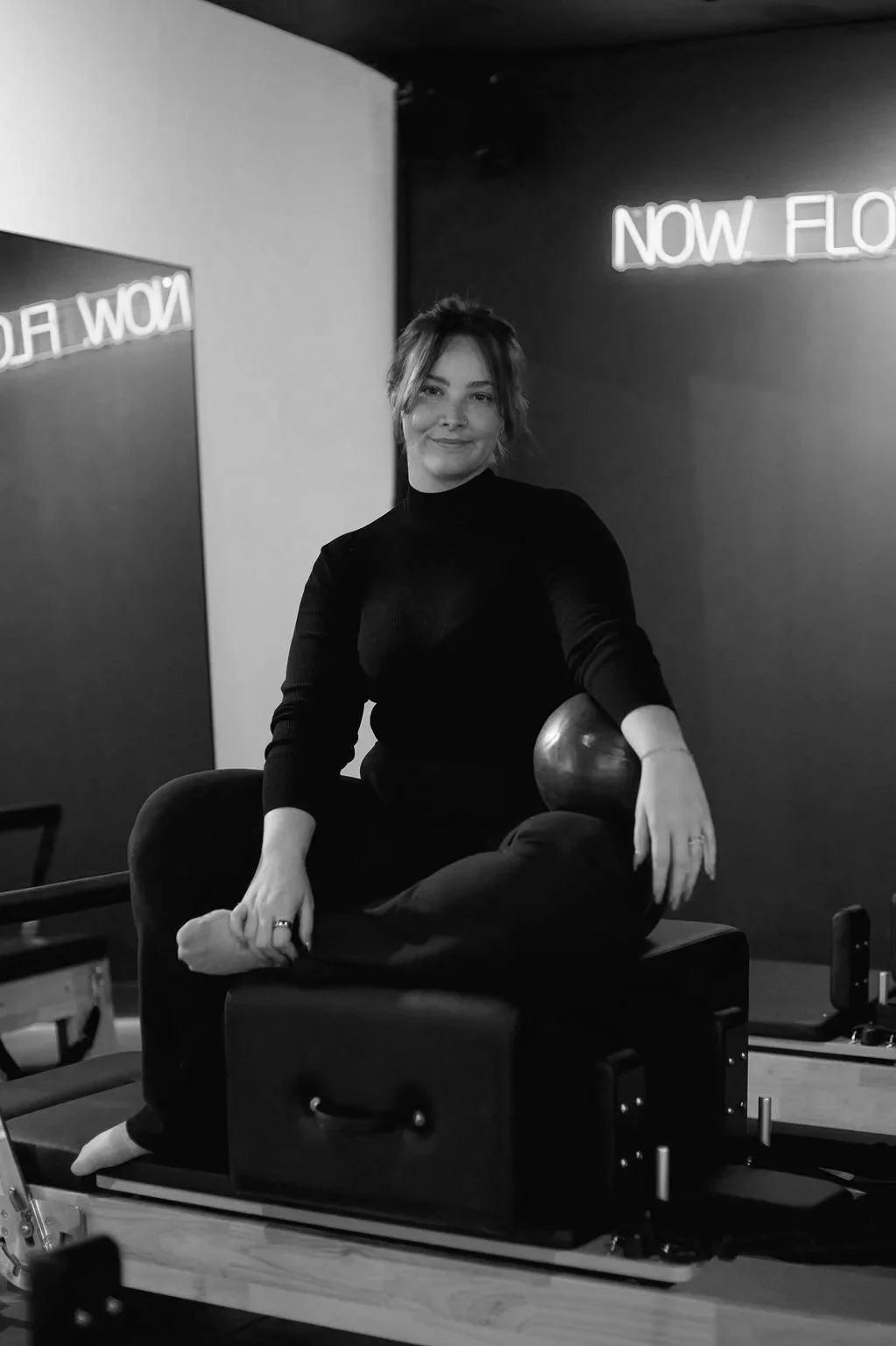 A woman with short hair, wearing a black long-sleeve top, sitting on a Pilates reformer machine in a fitness studio with neon signs reading 'NOW FLOW' and 'NOW FLOW' in the background.