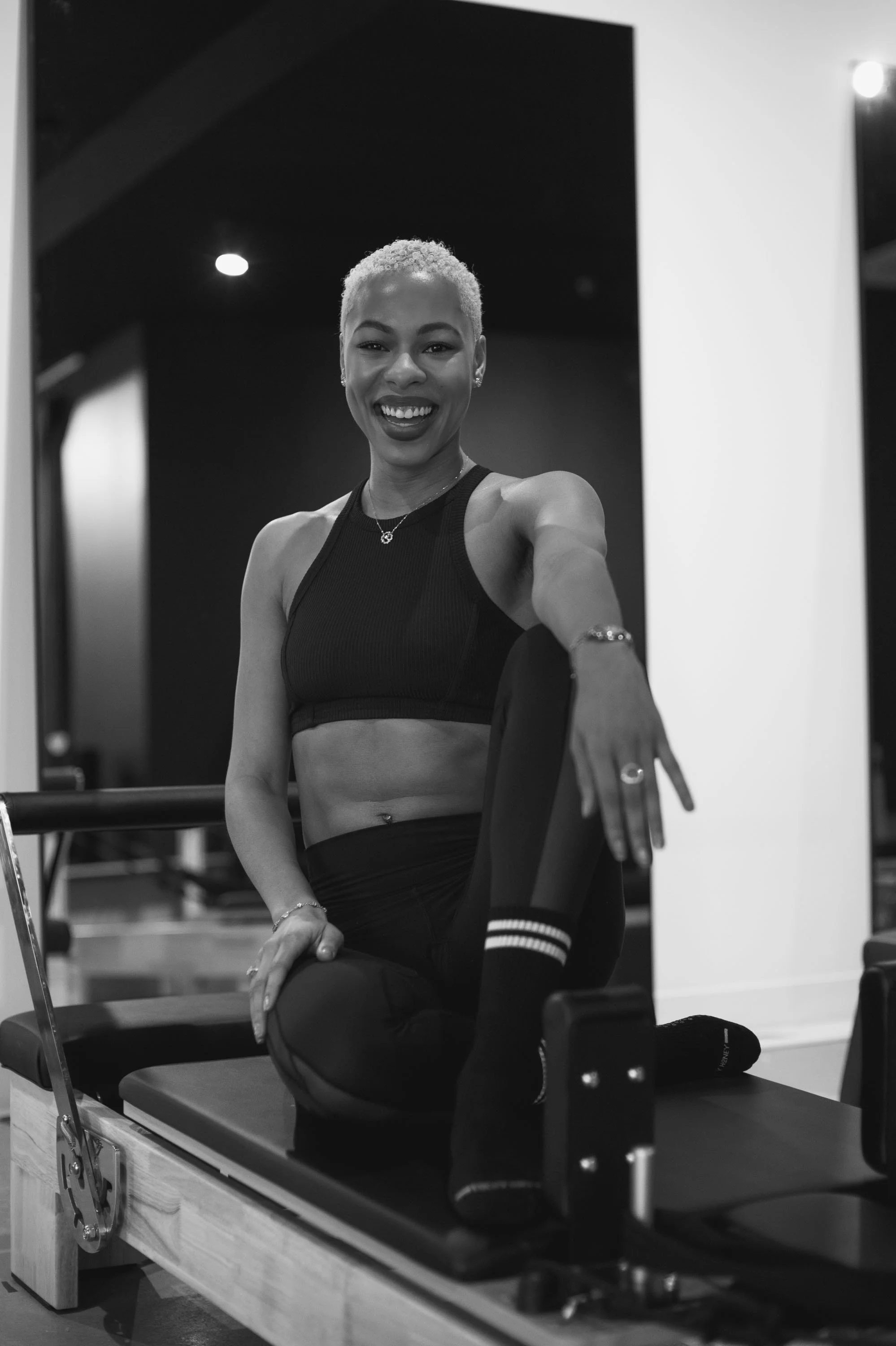 A woman with short platinum blonde hair smiling at the camera while kneeling on a Pilates reformer machine in a gym, wearing a black athletic top, black tights with stripes, and socks.