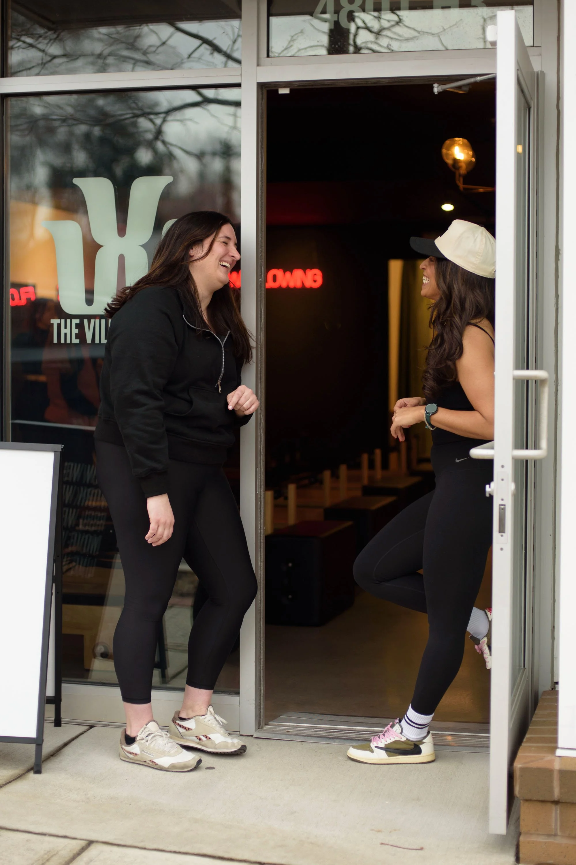 Two women are laughing and smiling while standing in front of a gym entrance, wearing athletic clothing, with the gym's logo and neon sign visible inside.