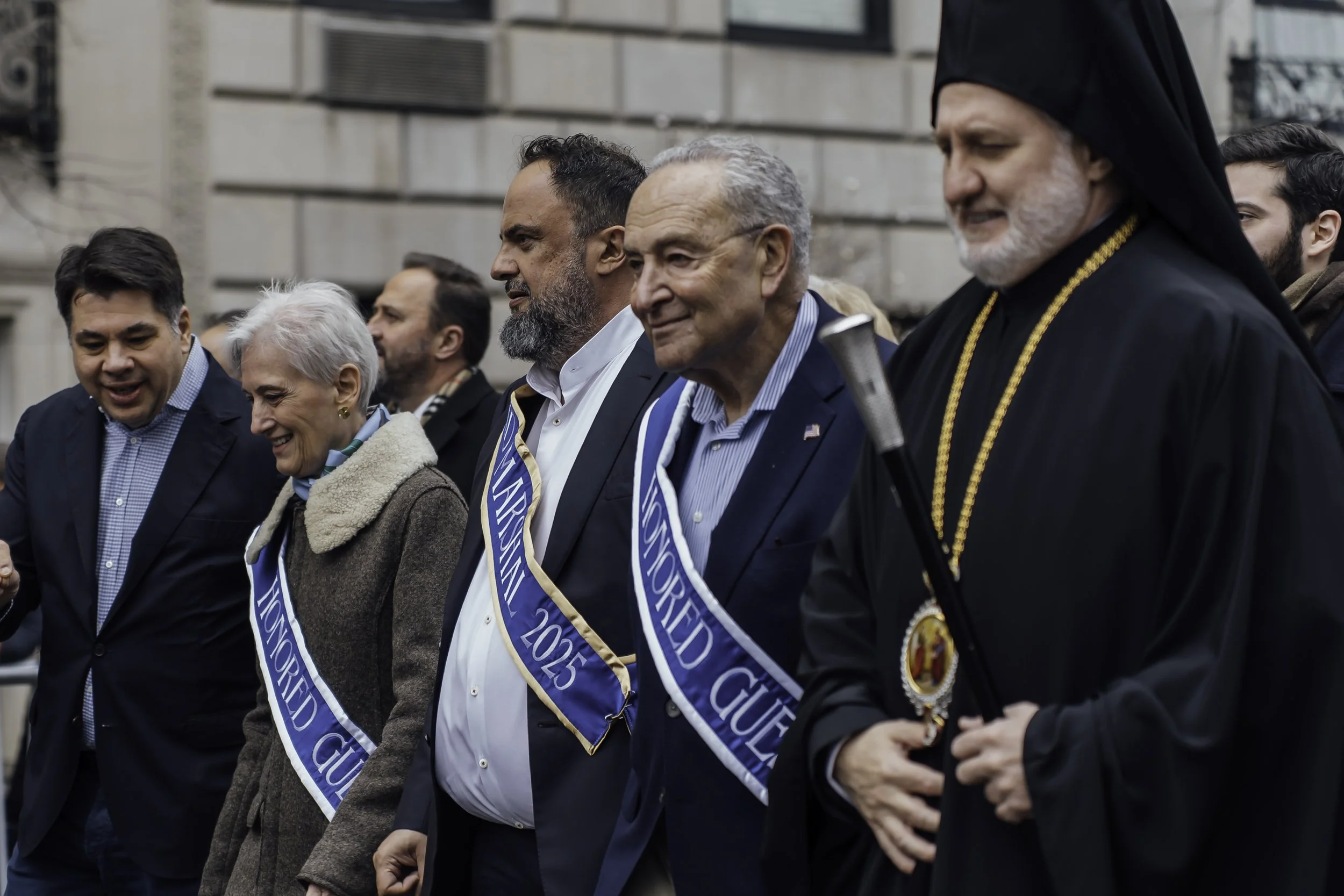 Line of people walking outdoors, wearing sashes that read "Honored Guest 2015," some smiling, in front of a building with stone walls.