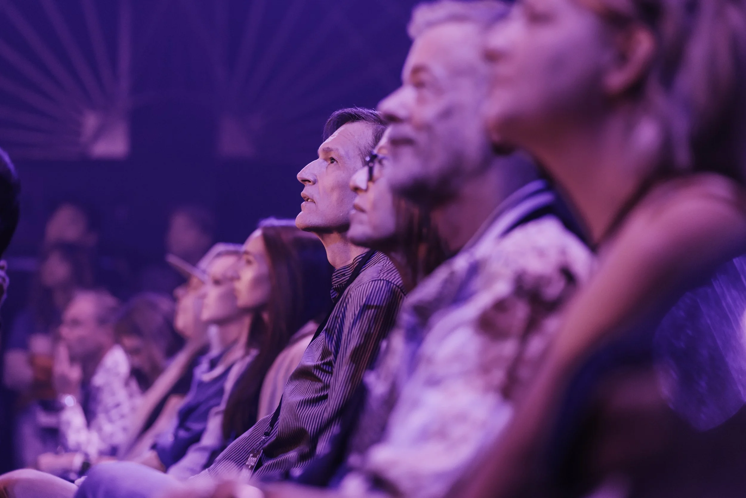 Audience of diverse individuals seated and attentively watching a presentation or event, illuminated by purple and blue lighting.