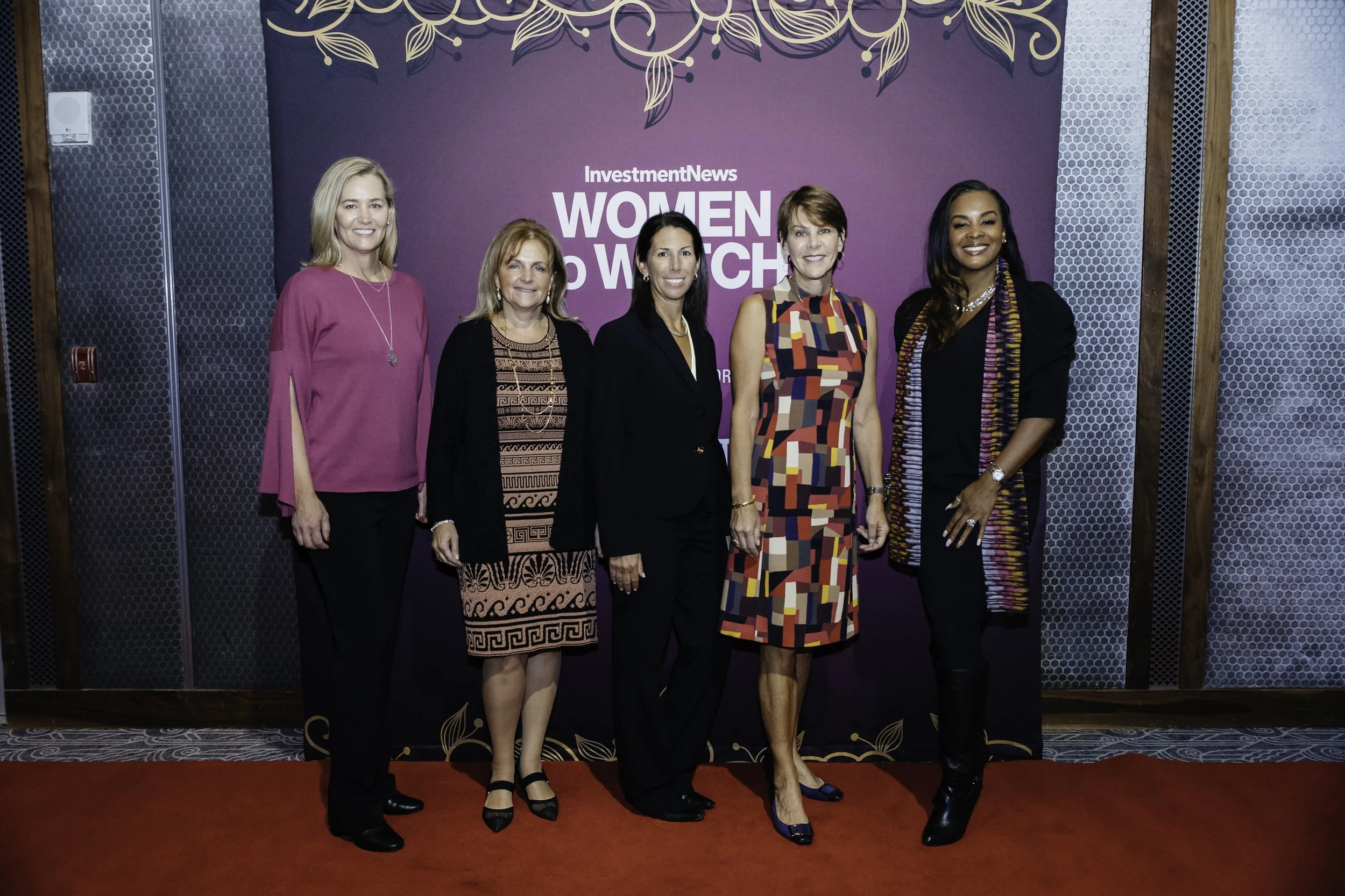 Six women standing on a red carpet in front of a purple backdrop with gold floral designs, posing for a photo at the InvestmentNews Women to Watch event.