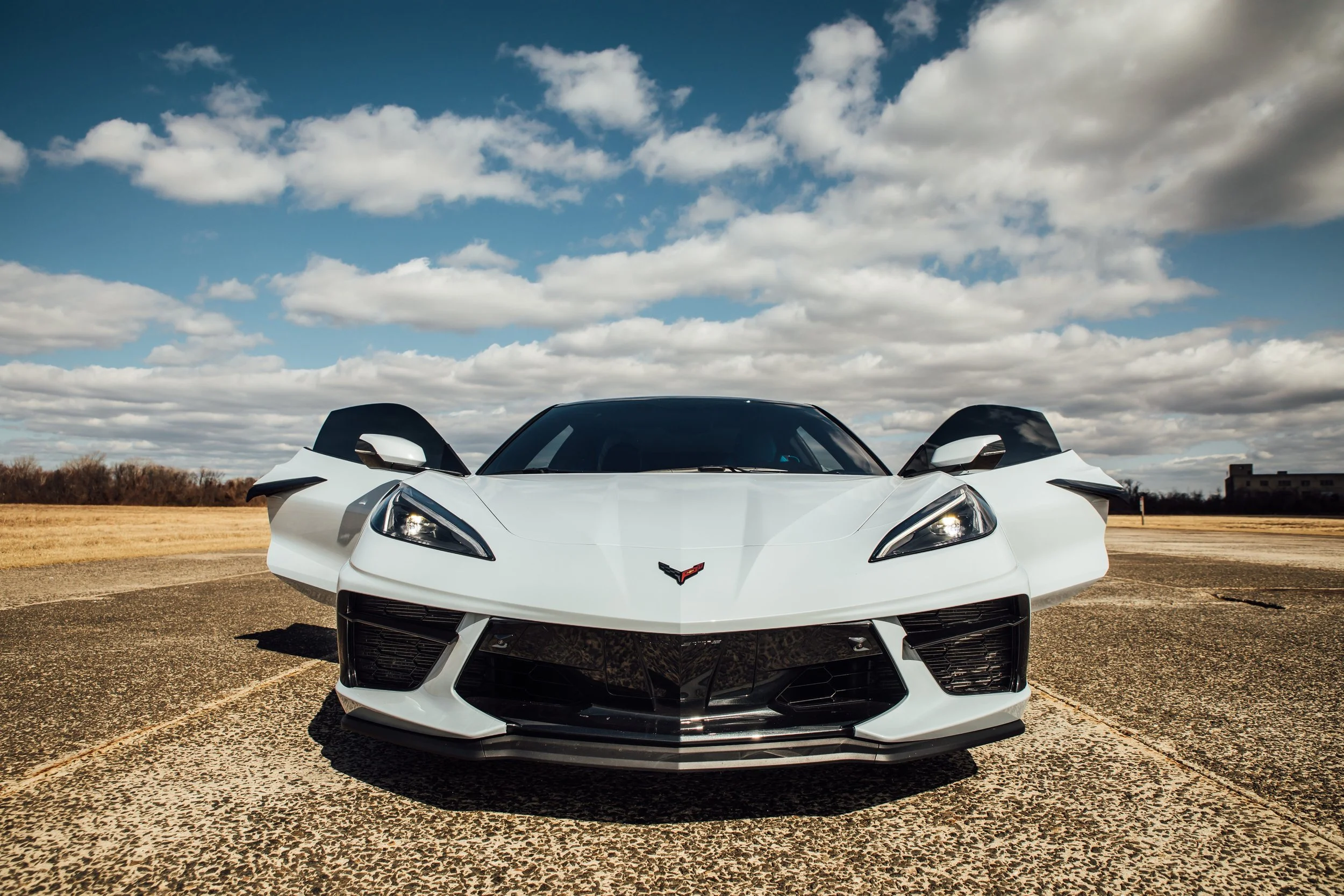 A white sports car with its doors open, parked on an empty lot under a partly cloudy sky.