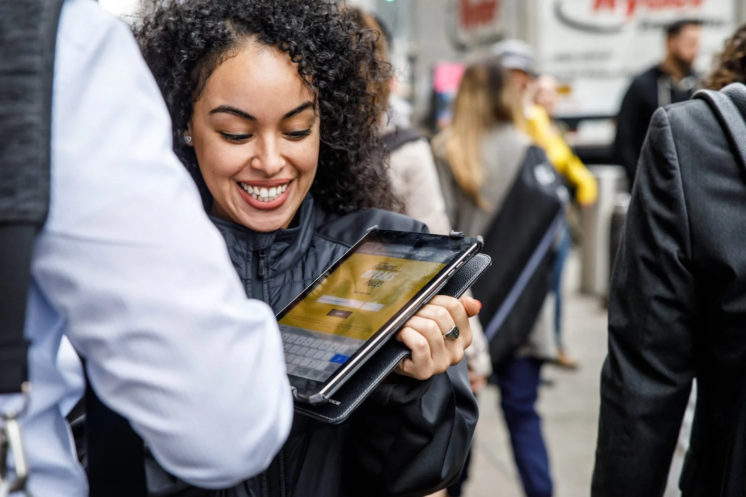 A woman with curly hair smiling at her tablet device while standing in a crowd.