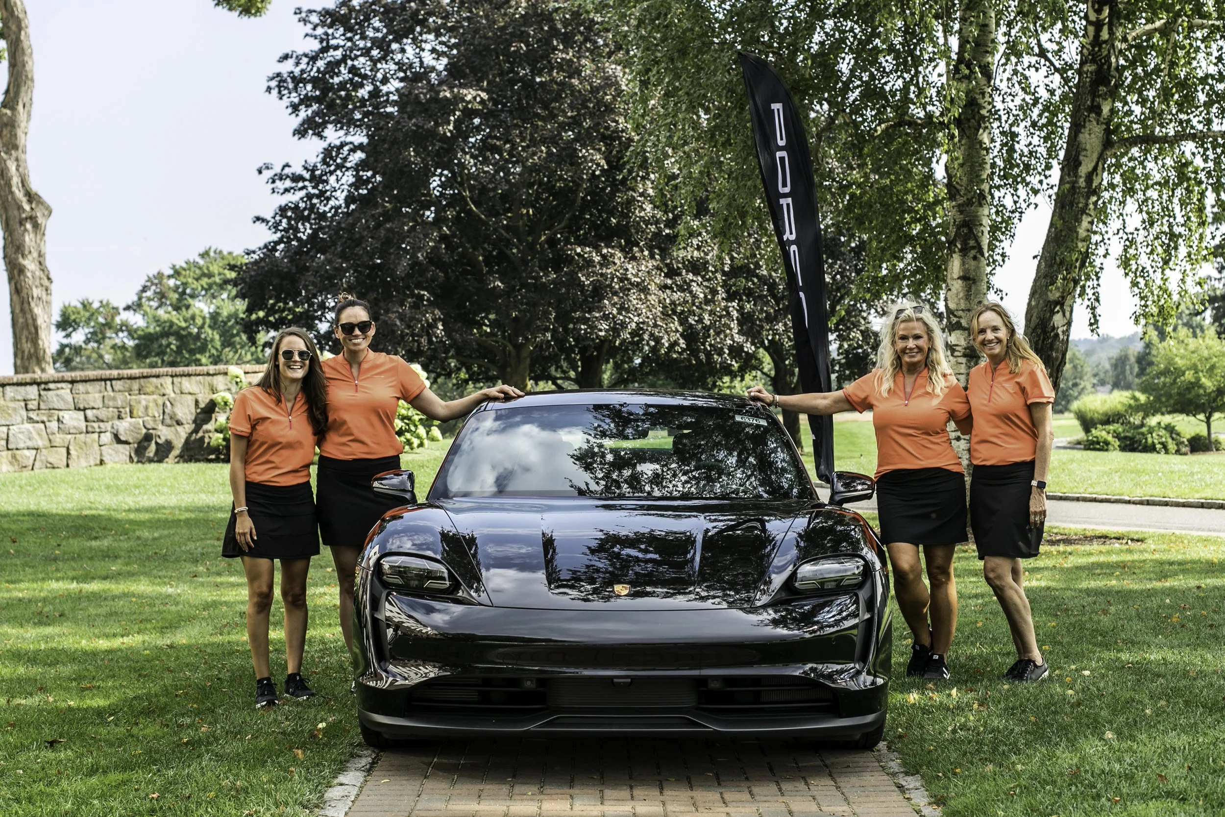 Four women in matching orange shirts and black skirts standing around a black sports car in a park with trees in the background.