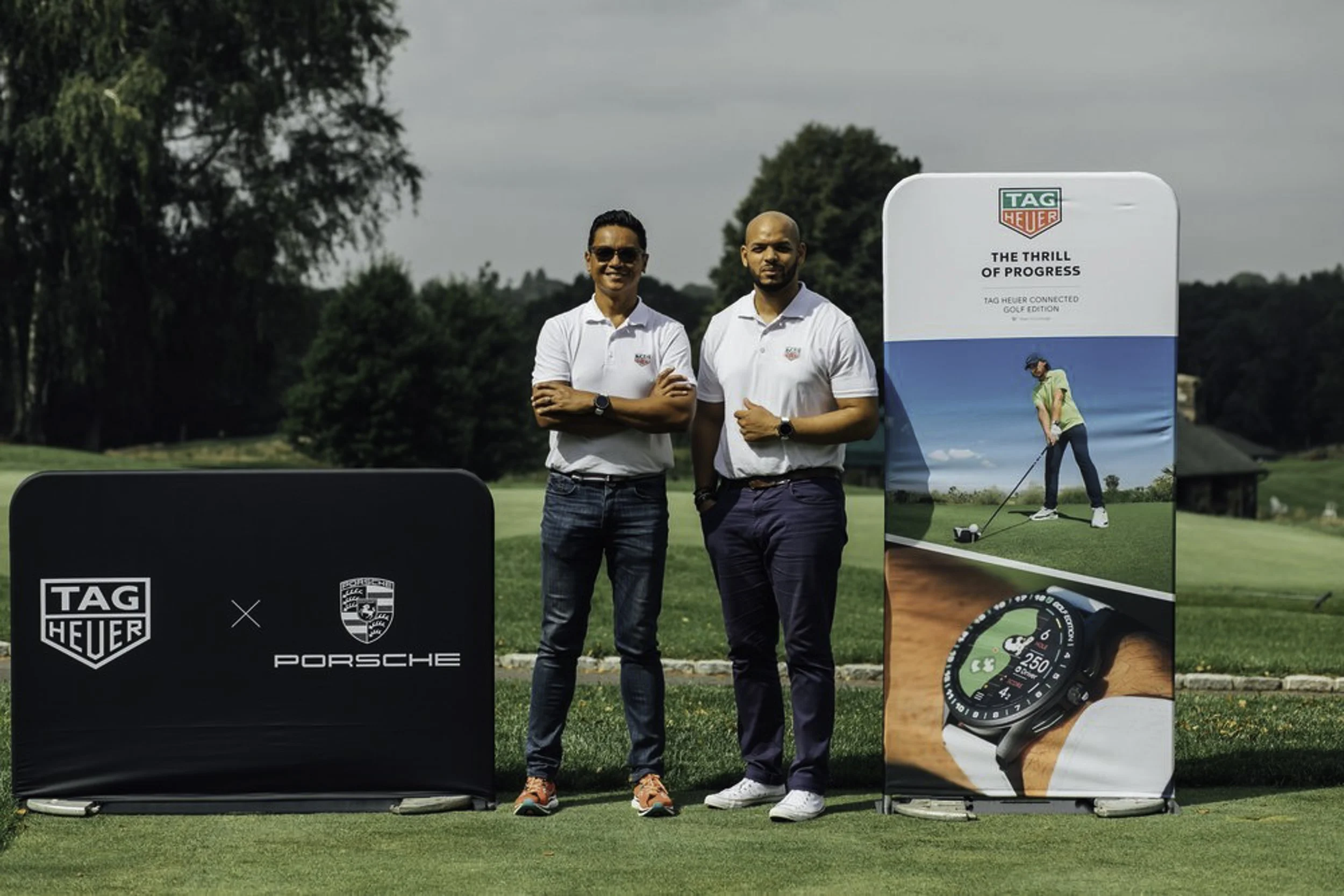 Two men standing on a golf course near promotional displays for TAG Heuer and Porsche, with trees and a building in the background.