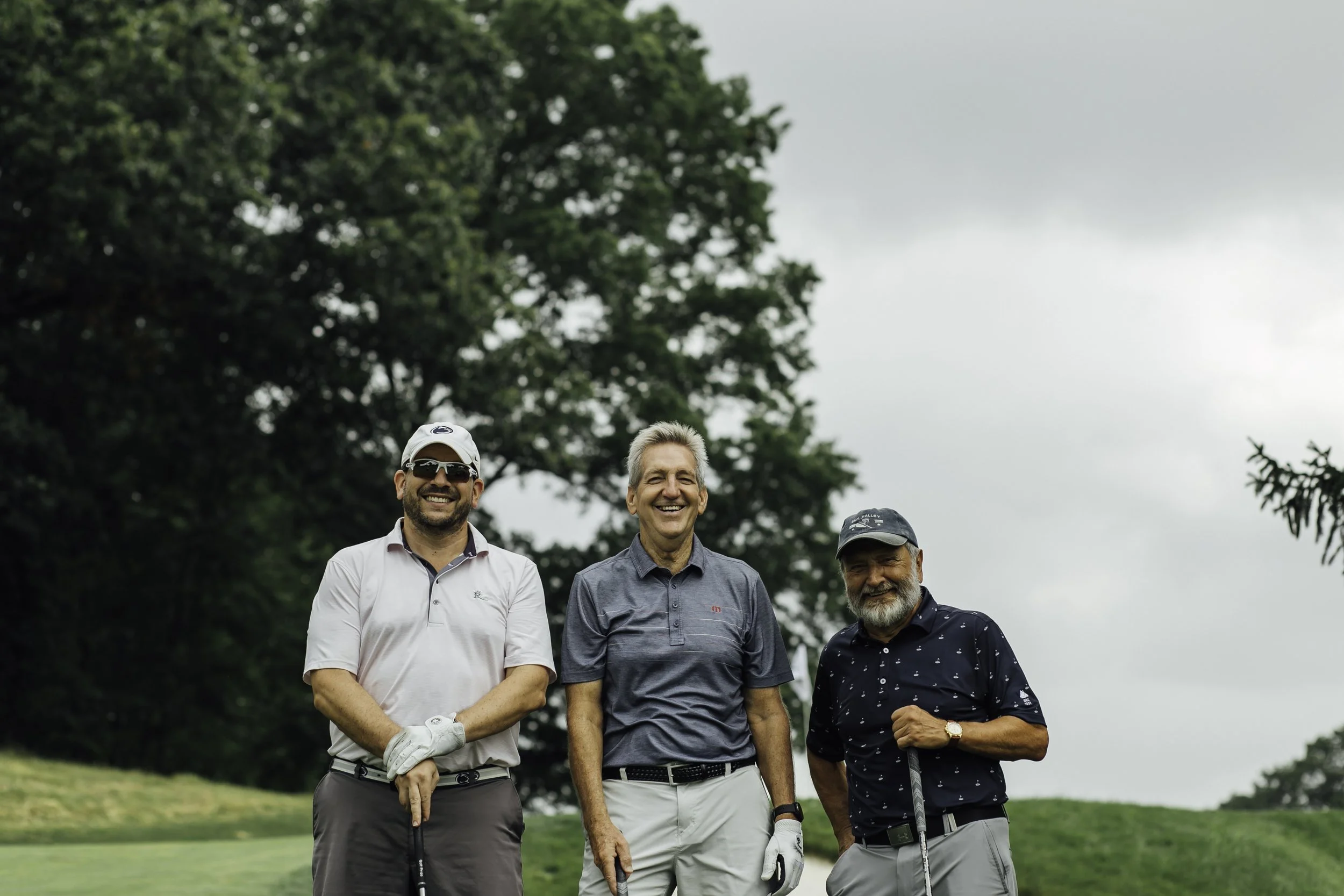 Three men standing on a golf course, smiling, with trees in the background, two holding golf clubs.