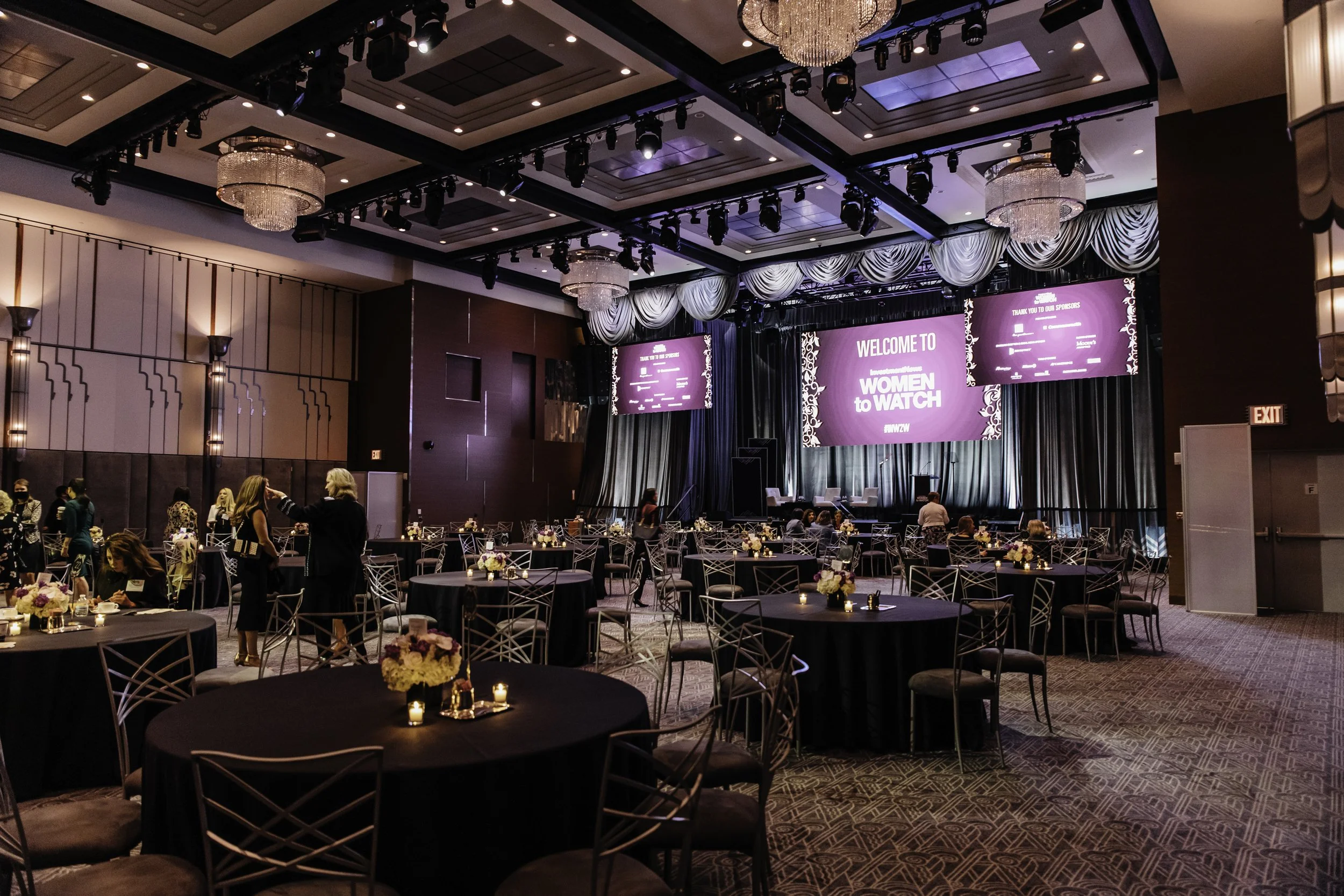 Elegant event venue with round tables decorated with flower centerpieces and candles, a large stage with purple screens displaying Welcome message for 'Women to Watch,' chandeliers hanging from the ceiling, and people mingling.