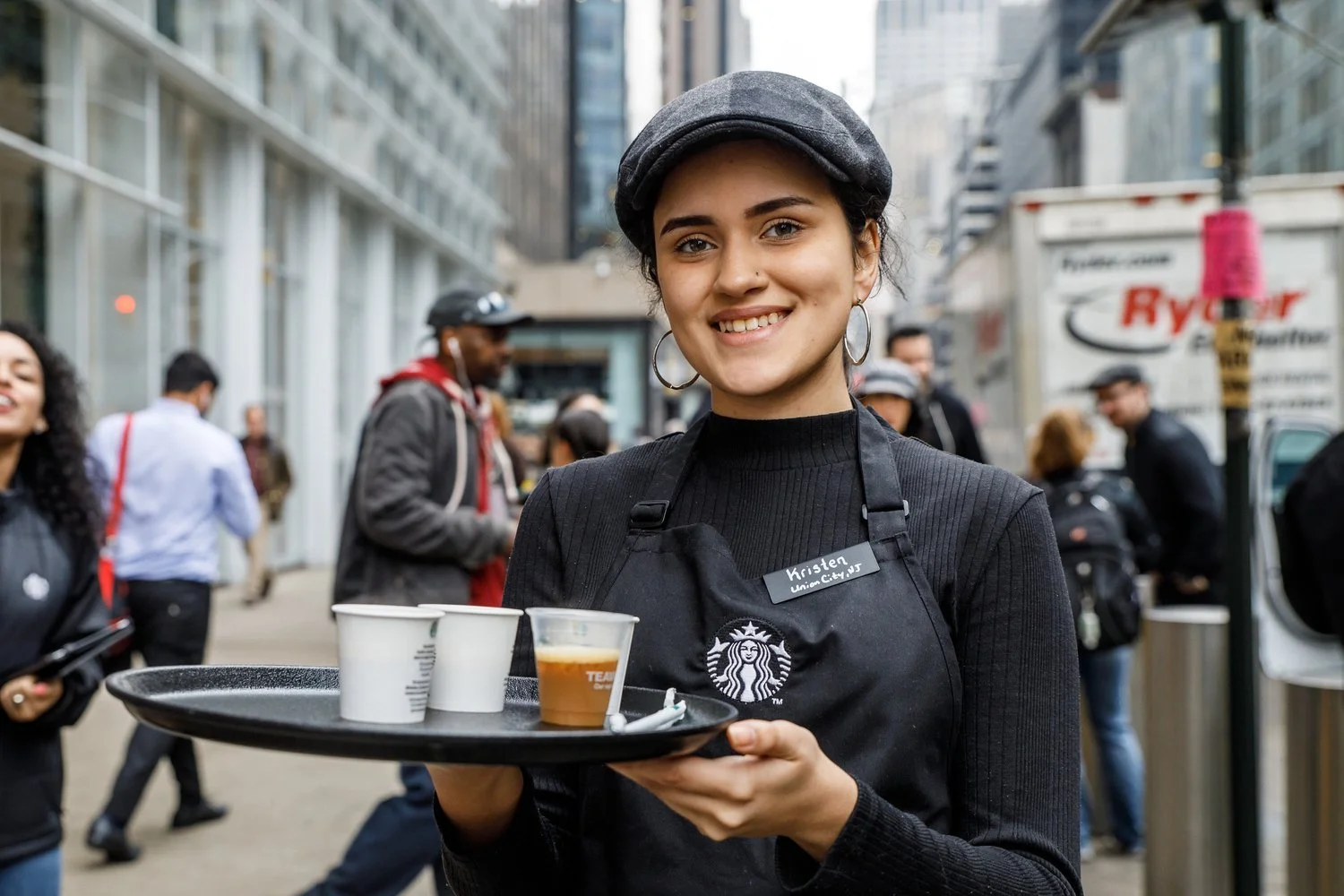 A young woman wearing a black Starbucks apron and a gray beret, smiling and holding a tray with cups, standing on a busy city street.