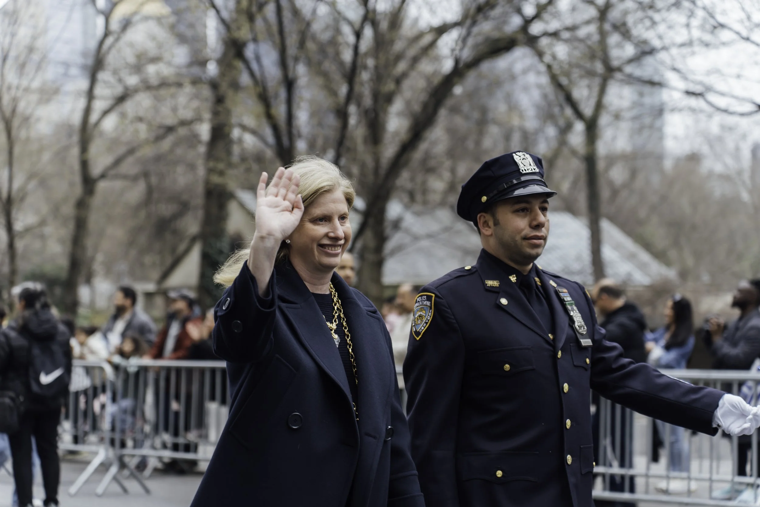 A woman in a navy blazer waving with a smile, walking beside a male police officer in uniform, during an outdoor public event with people and trees in the background.