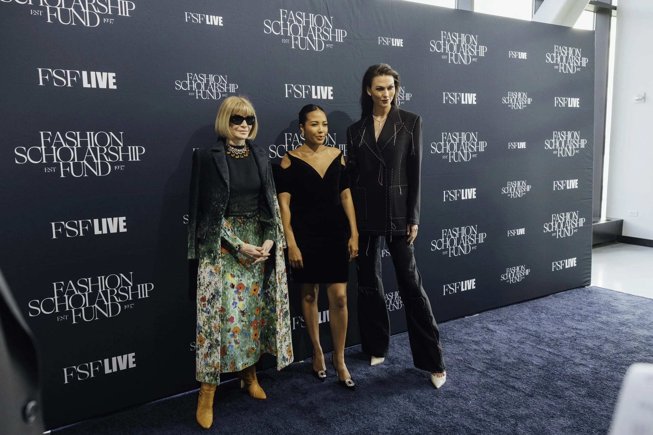 Three women standing in front of a dark backdrop with white text reading "Fashion Scholarship Fund EST 1957" and "FSF LIVE". The woman on the left wears sunglasses, a sparkly black blazer, a floral skirt, and tan boots. The woman in the middle wears a black dress with shoulder cutouts and high heels. The woman on the right wears a black suit with white stitching and white shoes.