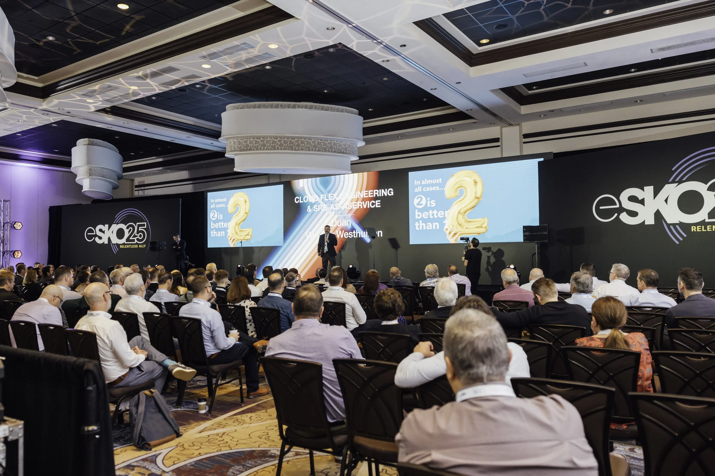Conference event with a large audience watching a speaker on stage at e-skoll 25th anniversary event. The stage has large screens displaying a presentation with the text 'In almost all cases, 2 is better than 1' and a logo for e-skoll on a black background.