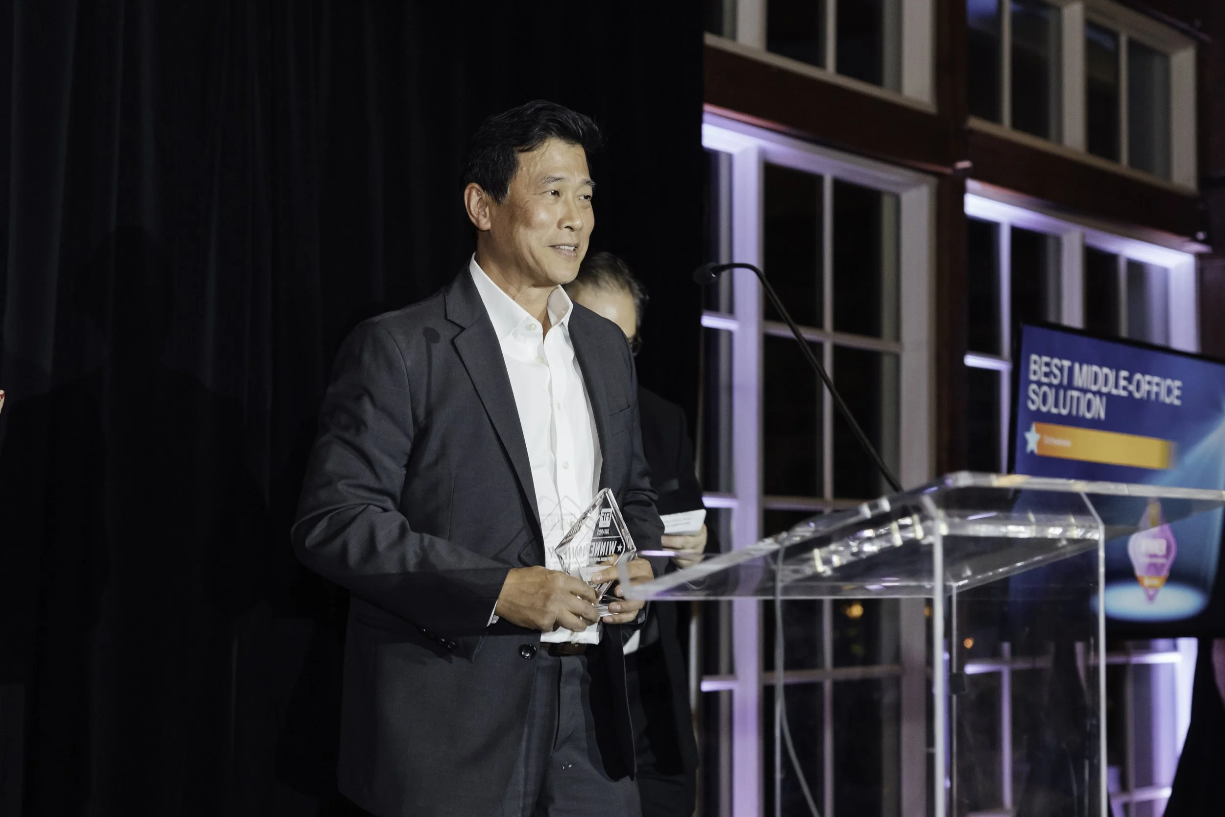 Man in a suit holding an award on stage at an event with a sign that says 'Best Middle Office Solution' in the background.