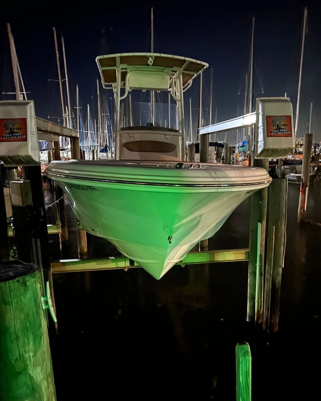 A motorboat docked at a marina during nighttime, illuminated by green lights with multiple sailboats moored in the background.