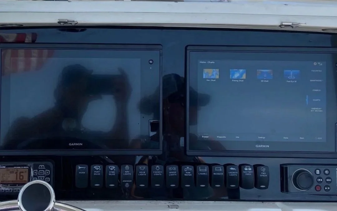 Two Garmin marine navigation screens mounted on a boat dashboard, with reflection of two people and the sky visible on the screens.