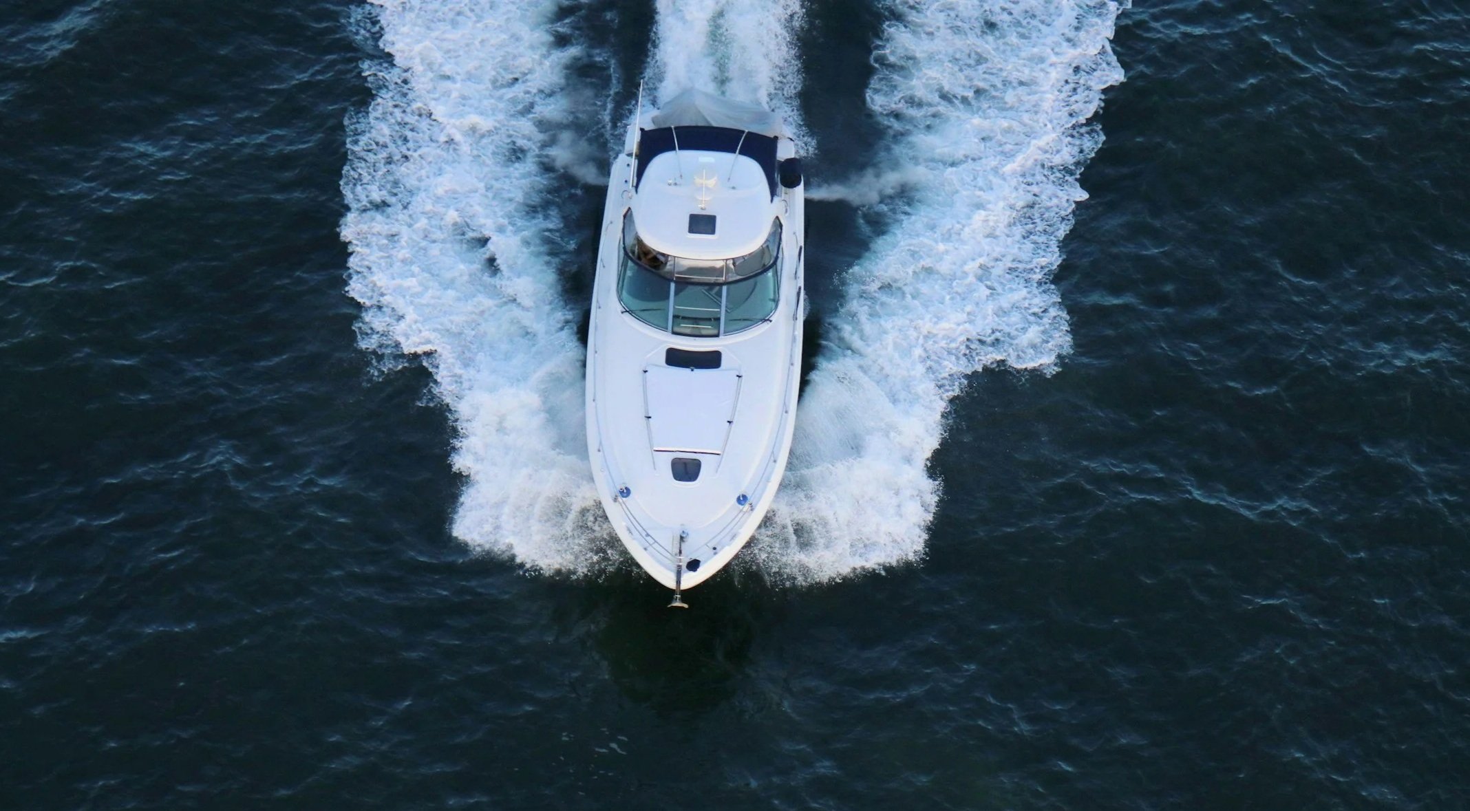 An aerial view of a white yacht moving through dark blue water, creating a wake behind it.