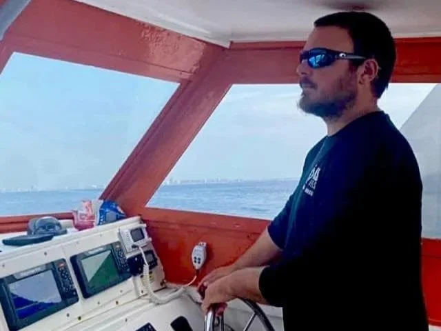 A man wearing reflective sunglasses and a dark blue shirt standing on a boat's bridge, with a view of the ocean and blue sky in the background.
