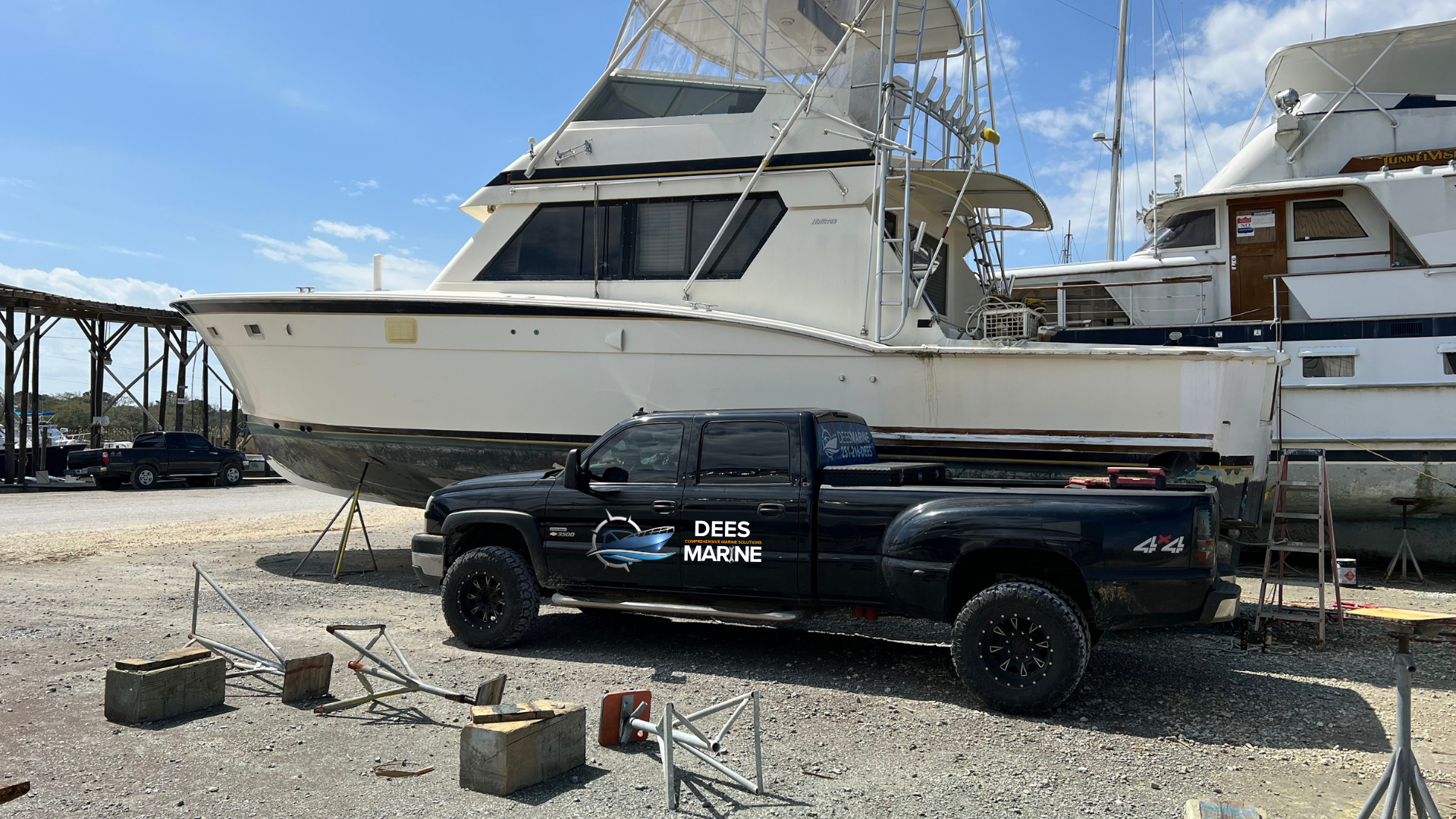 A large white boat on a trailer in a boatyard next to a black pickup truck with 'DEES MARINE' printed on the side. The boatyard has a gravel surface, with some debris and concrete blocks around, and other boats and pickup trucks in the background under a partly cloudy sky.